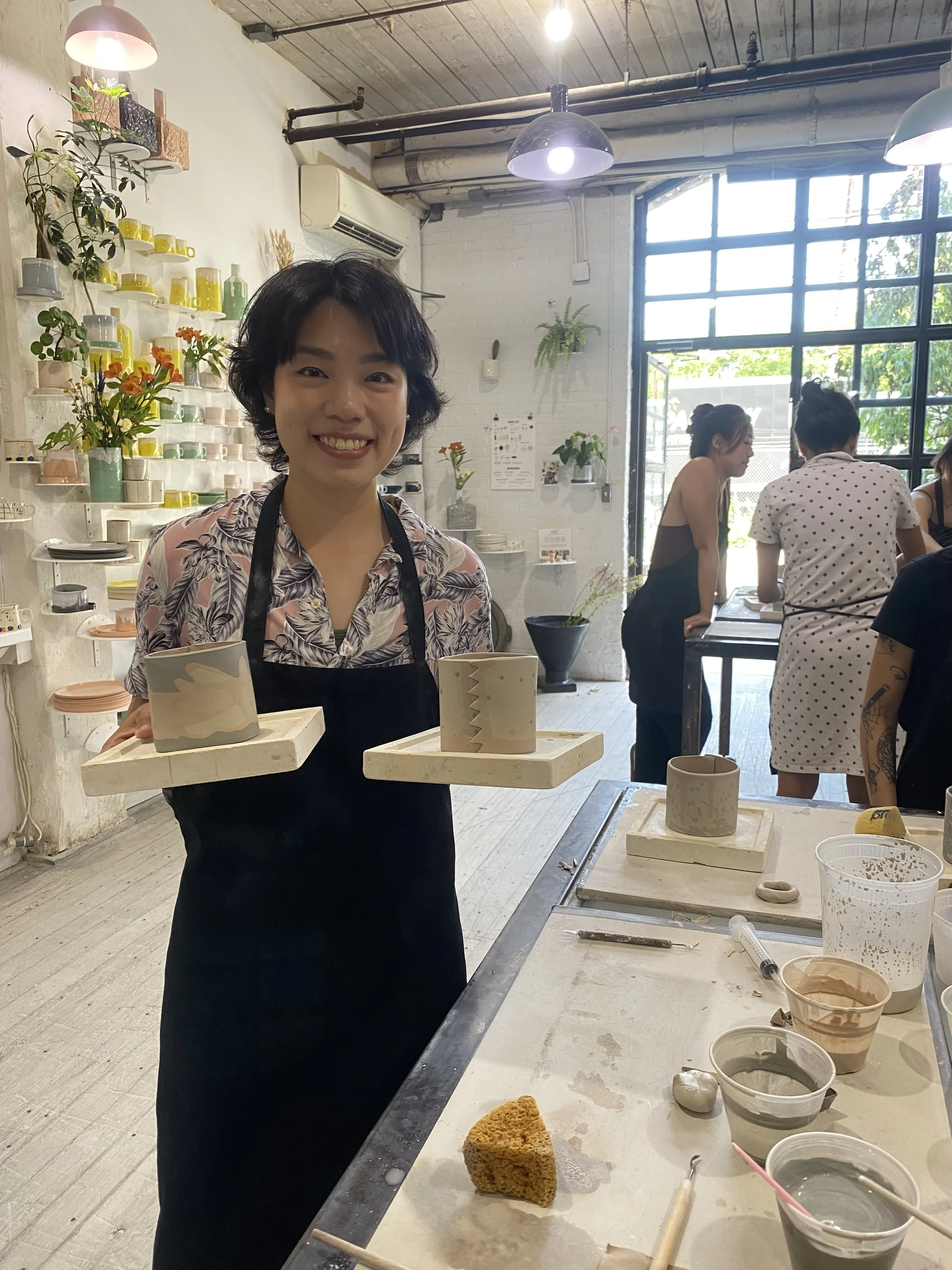 Beginner student smiling and holding two handmade ceramic mugs created during a handbuilding workshop at our Brooklyn pottery studio.