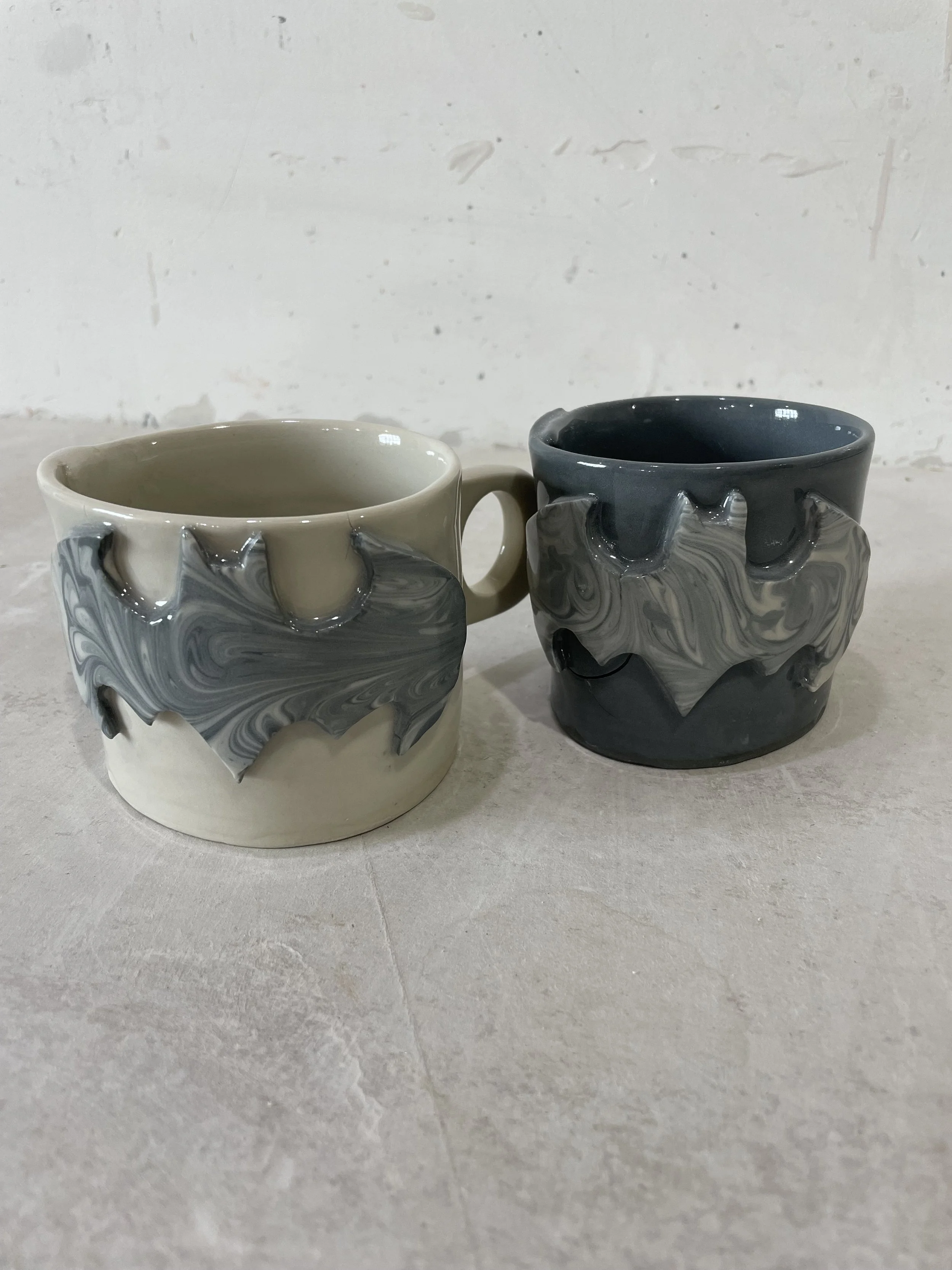 Two ceramic mugs with swirling marble patterns, one in cream and gray tones and the other in black and gray tones, placed on a light-colored surface against a white textured background.