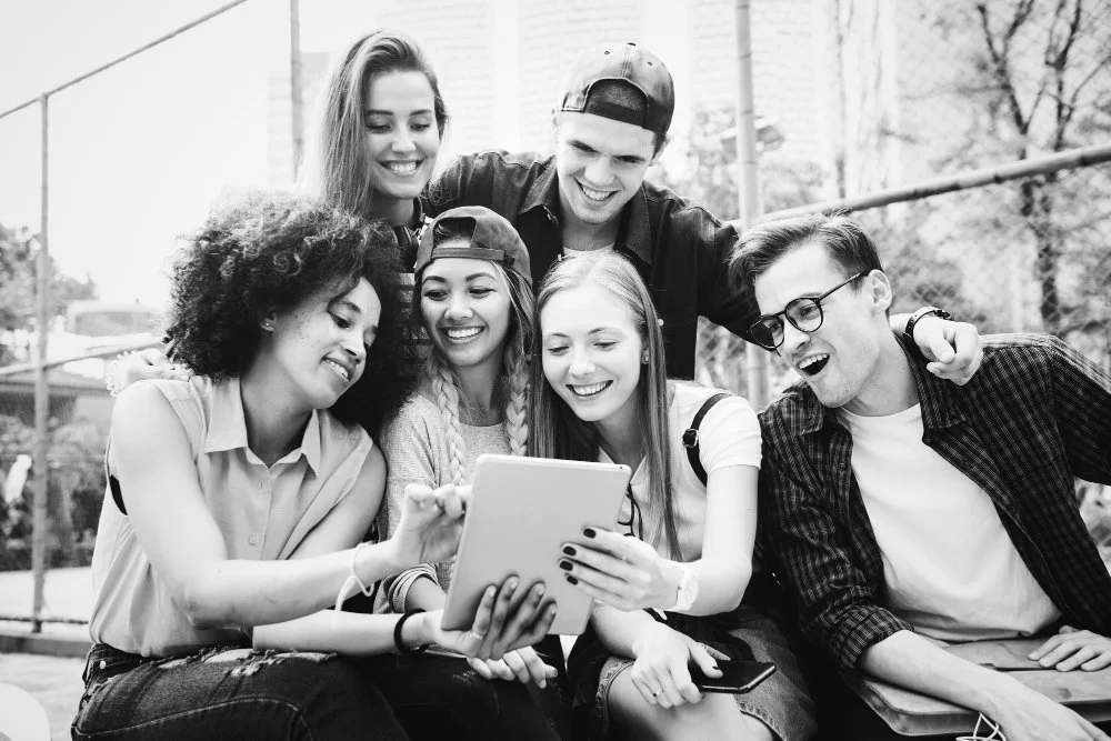 Six young people, four women and two men, gathered outdoors, smiling and looking at a tablet device.