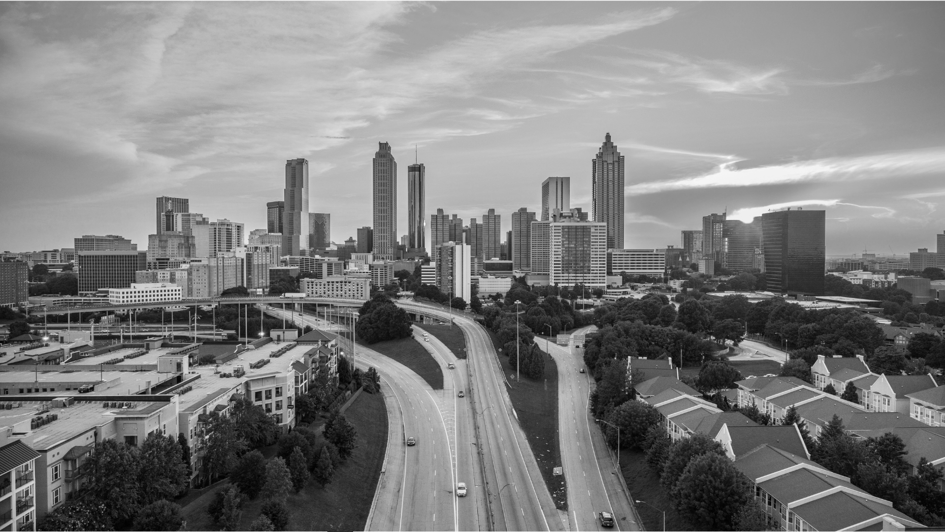 Black and white aerial view of a city skyline with high-rise buildings, residential area with townhouses, and a winding highway with a few cars.