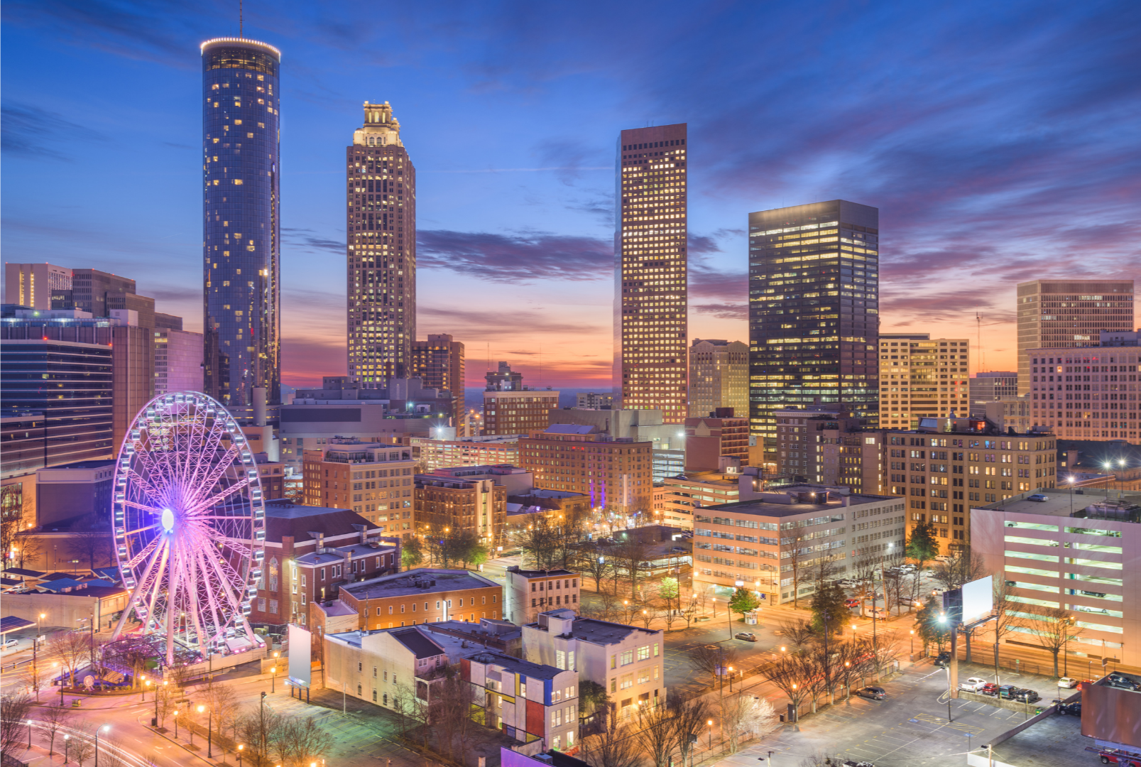 Nighttime city skyline with tall buildings, a lit-up Ferris wheel, and colorful sunset sky.