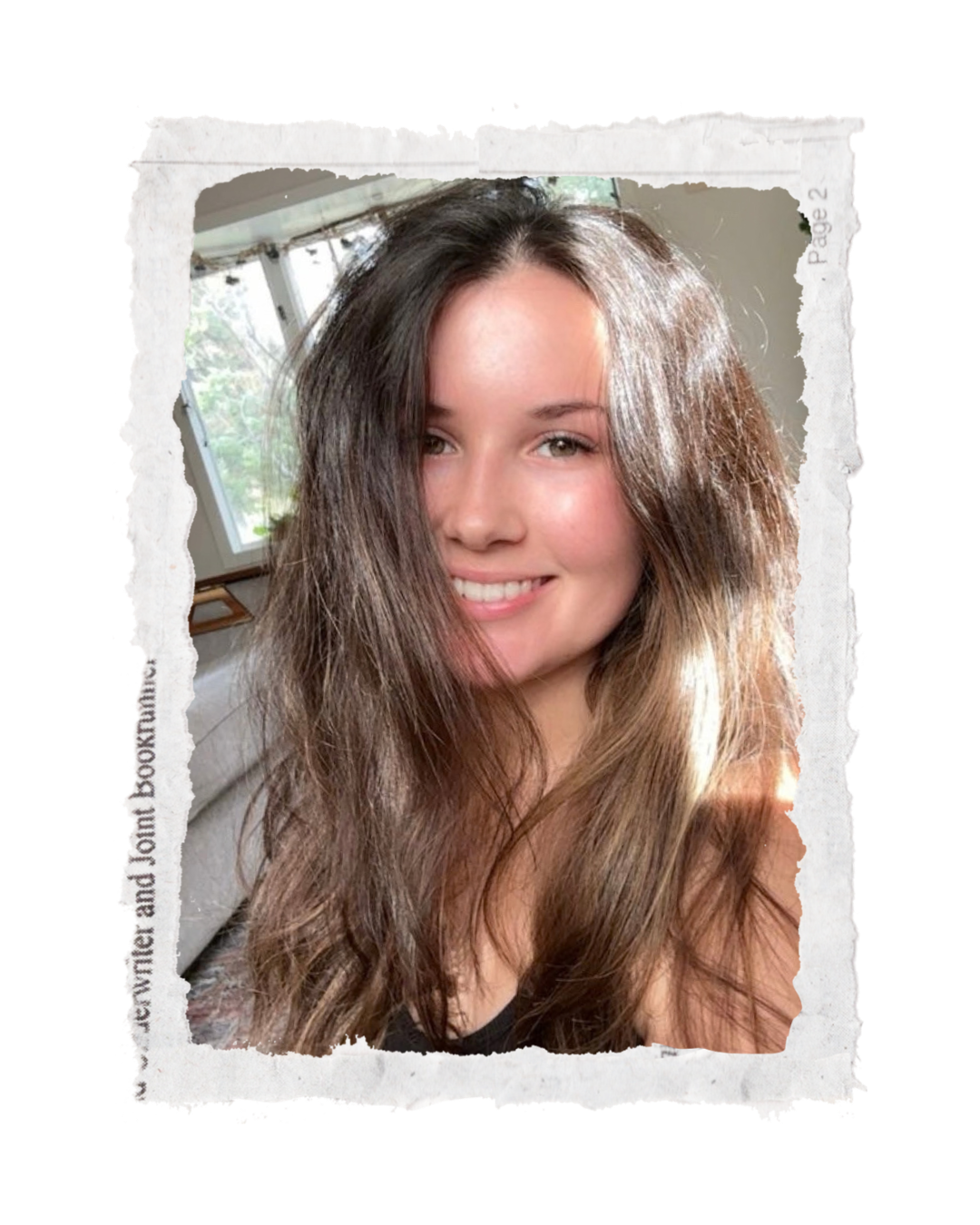 A young woman with long, wavy brown hair smiling at the camera, with sunlight illuminating her face, in front of a window with trees outside.