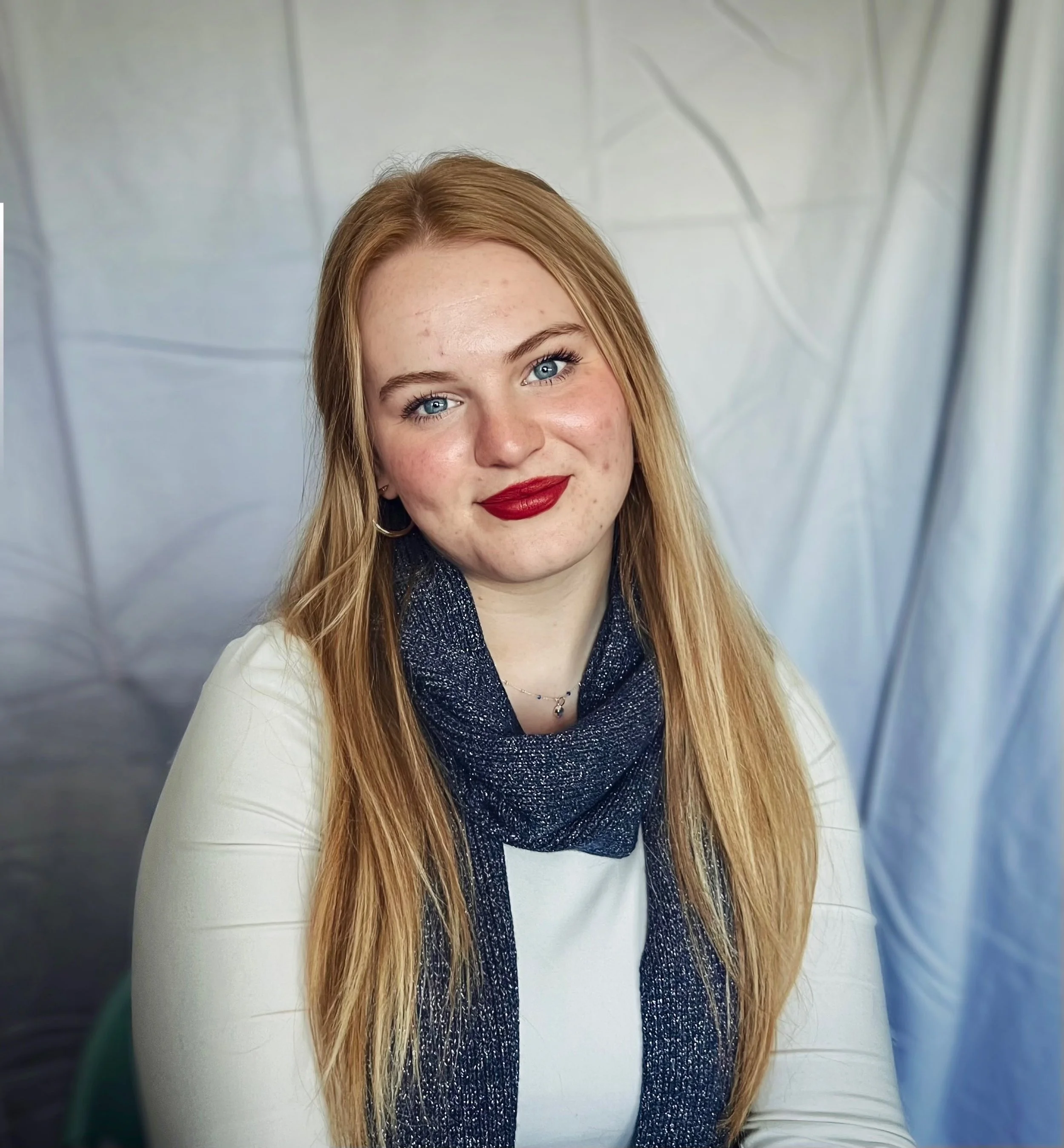 A young woman with long blonde hair, blue eyes, and red lipstick, wearing a white top with a dark blue scarf, smiling at the camera. The background features a metallic or plastic material with a few visible lines.
