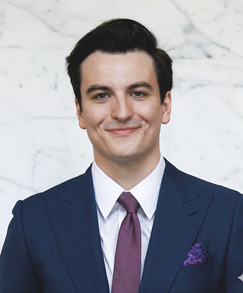 A young man with dark hair wearing a navy suit, white shirt, purple tie, and purple pocket square, smiling in front of a white marble background.