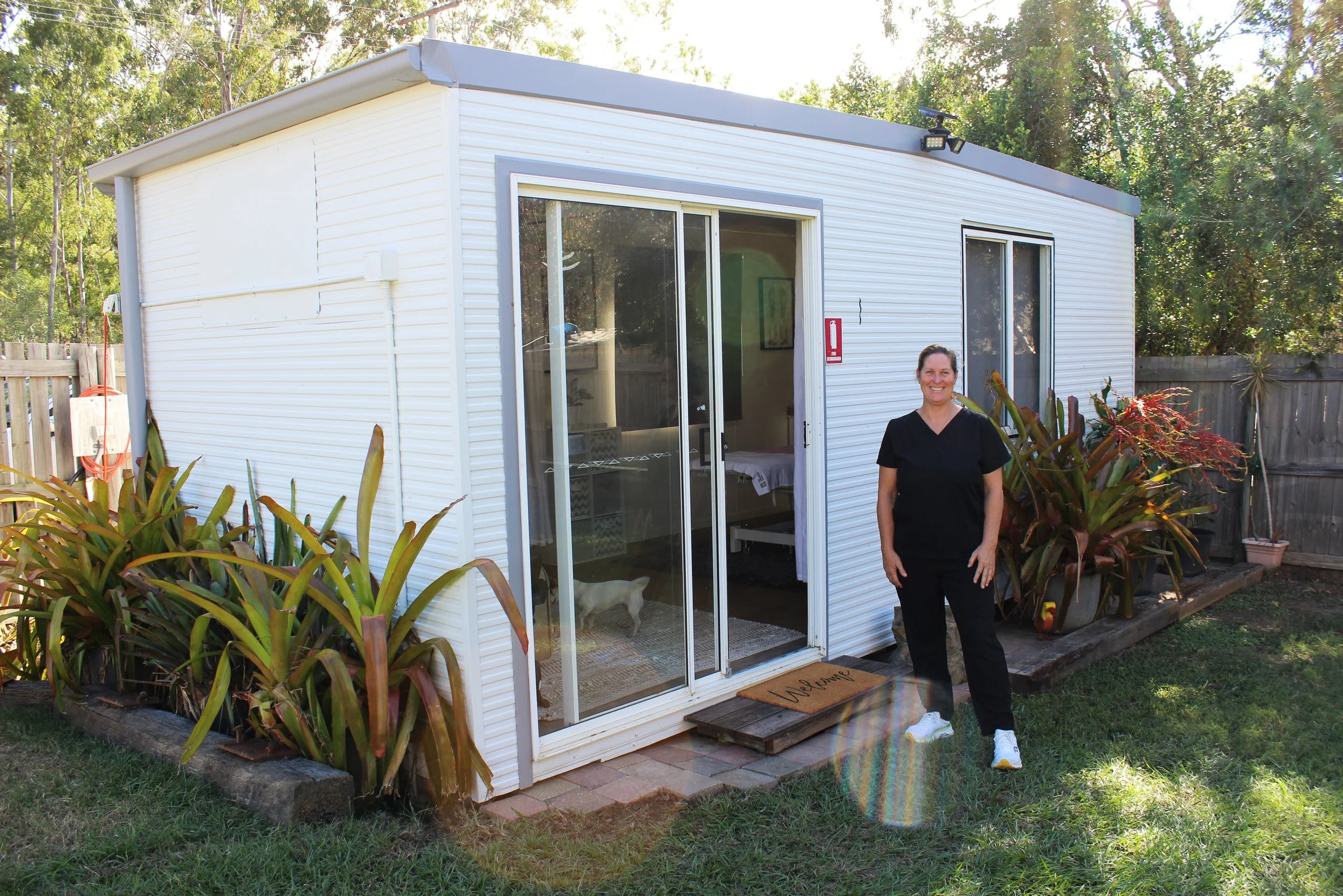 A remedial massage therapist in black scrubs standing outside a small white modular massage therapy room with an open sliding glass door, surrounded by garden plants, in a backyard with a wooden fence and trees in the background.