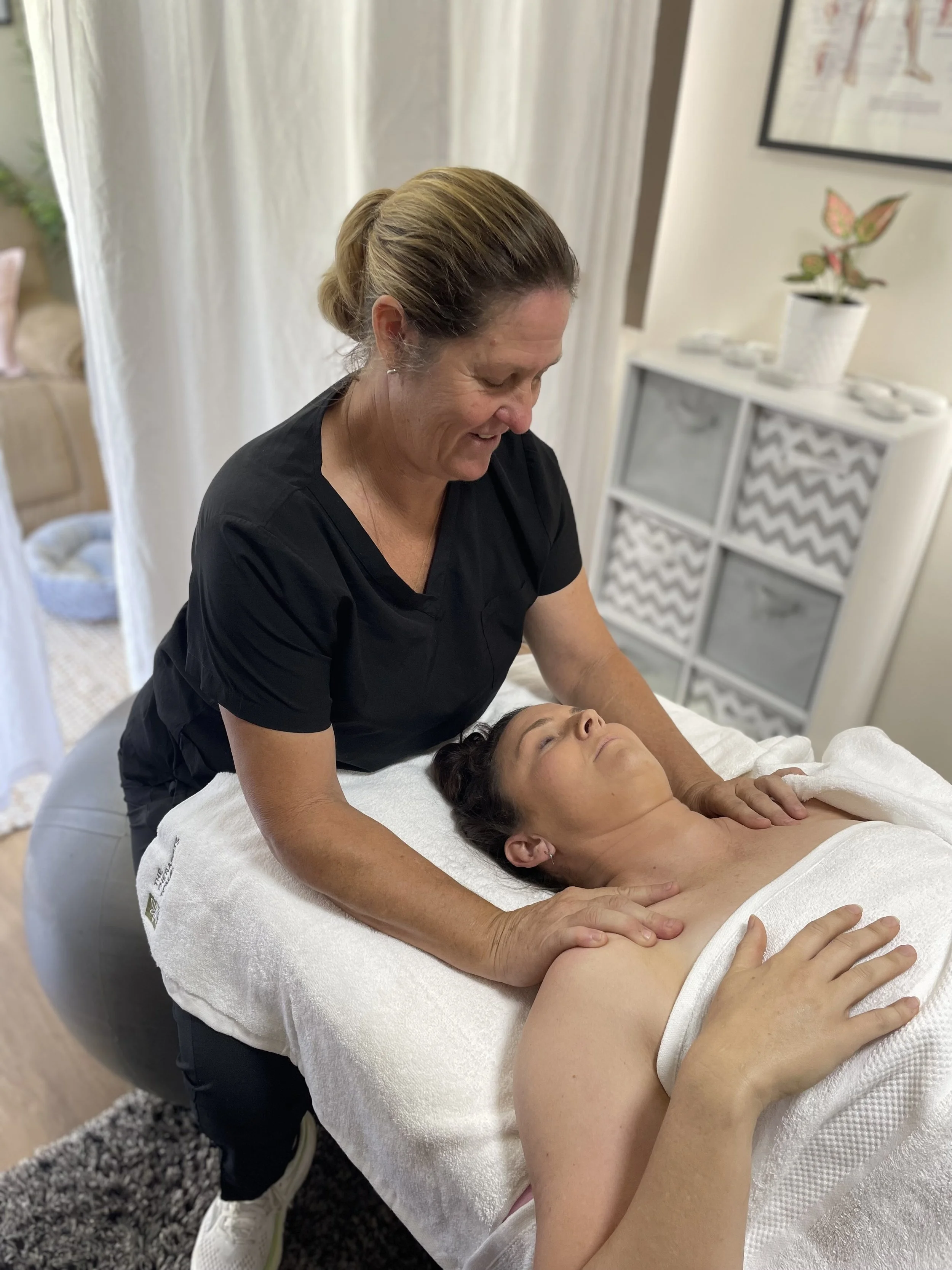 A woman lying on a massage table receiving a remedial massage while a therapist provides shoulder and neck massage in a massage therapy room.