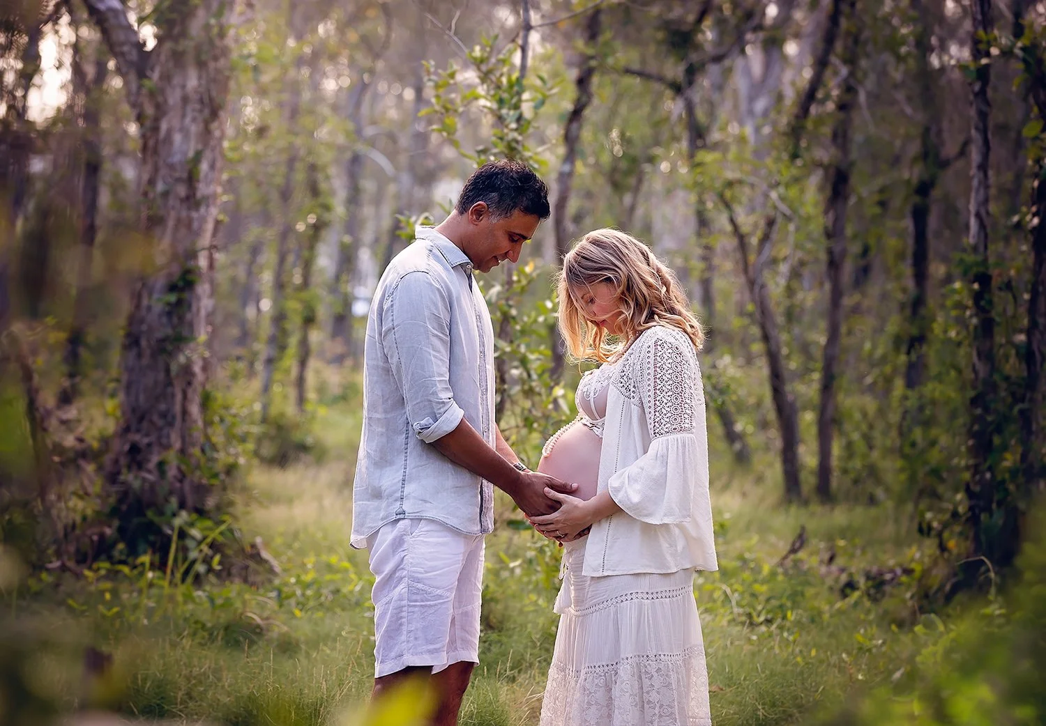 Pregnant woman and partner standing together in a forest, holding her belly.