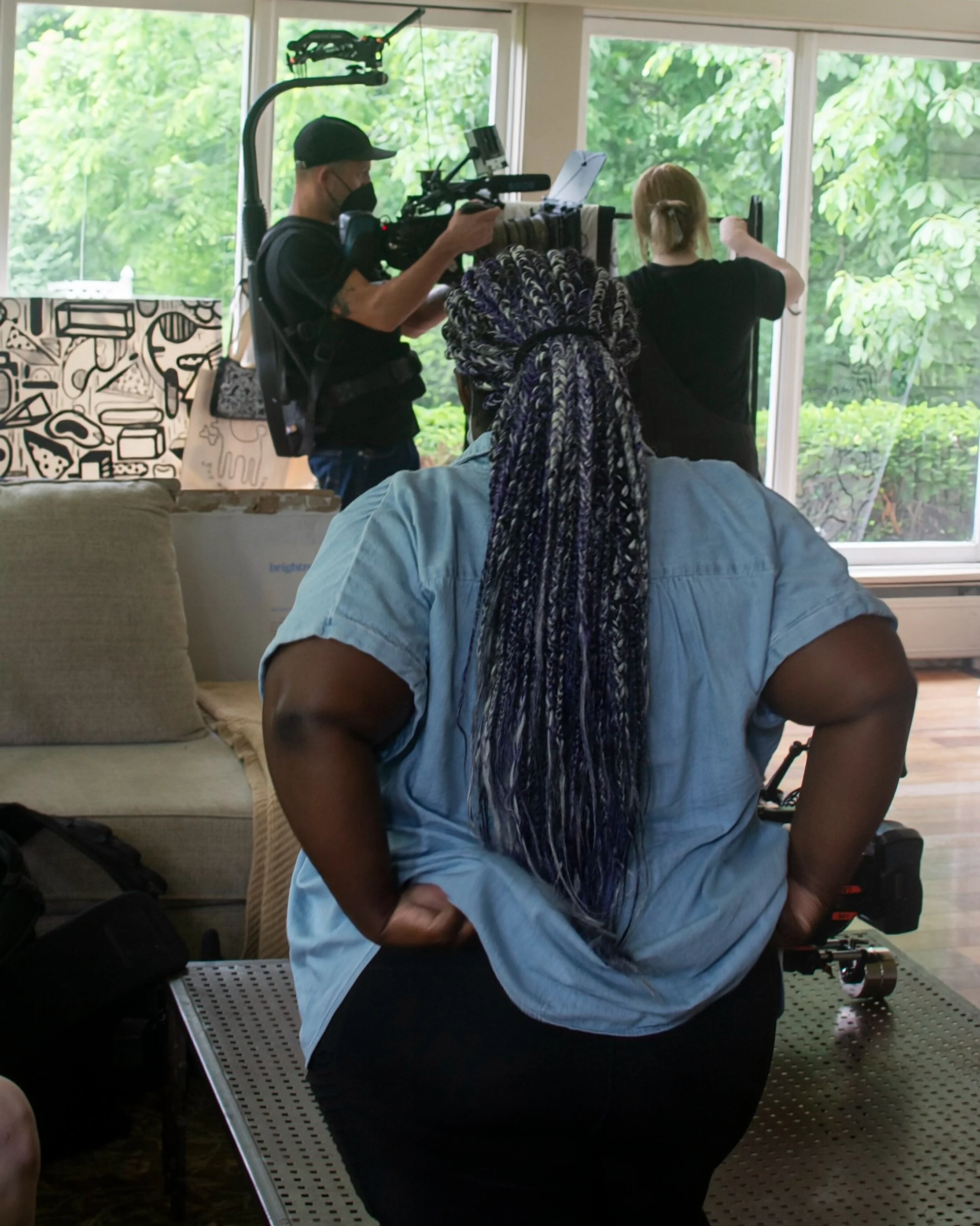 Efa's back is turned to us showing long braids pulled into a ponytail and a blue denim shirt. She is watching a cameraman film a woman in close proximity as she shows off original clothing designs in a spacious living room with full length windows.