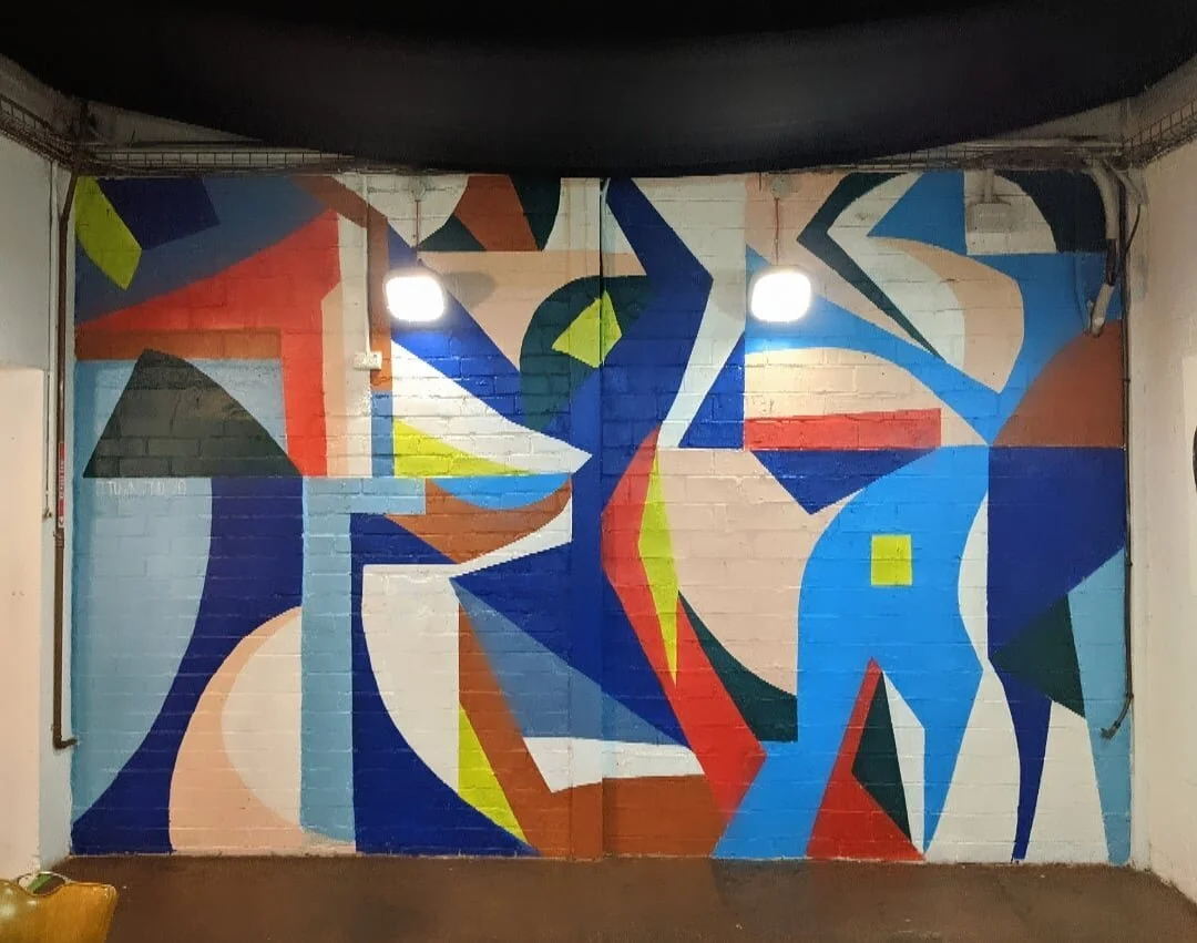Colorful abstract geometric mural painted on a brick wall, illuminated by two lights.