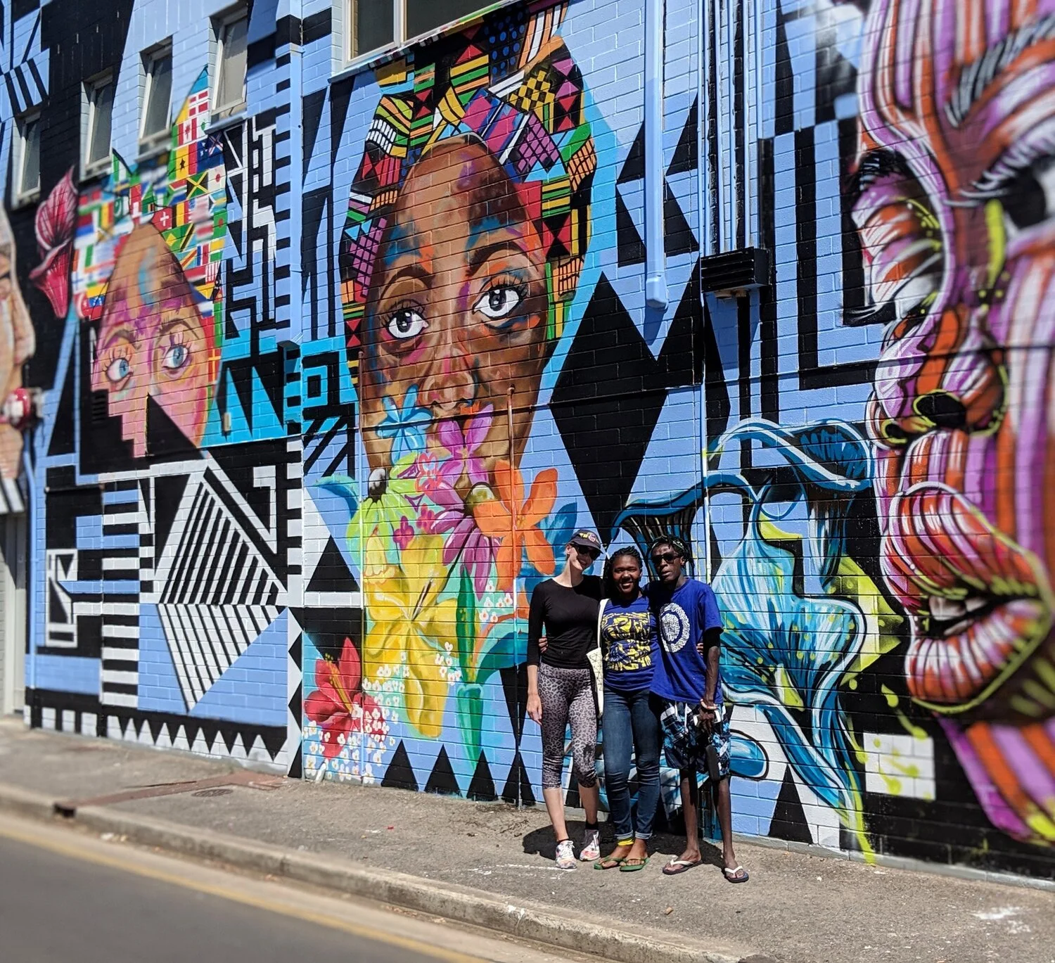 Three women standing in front of a vibrant mural on a brick wall featuring large portraits of women's faces, colorful flowers, and abstract geometric patterns.