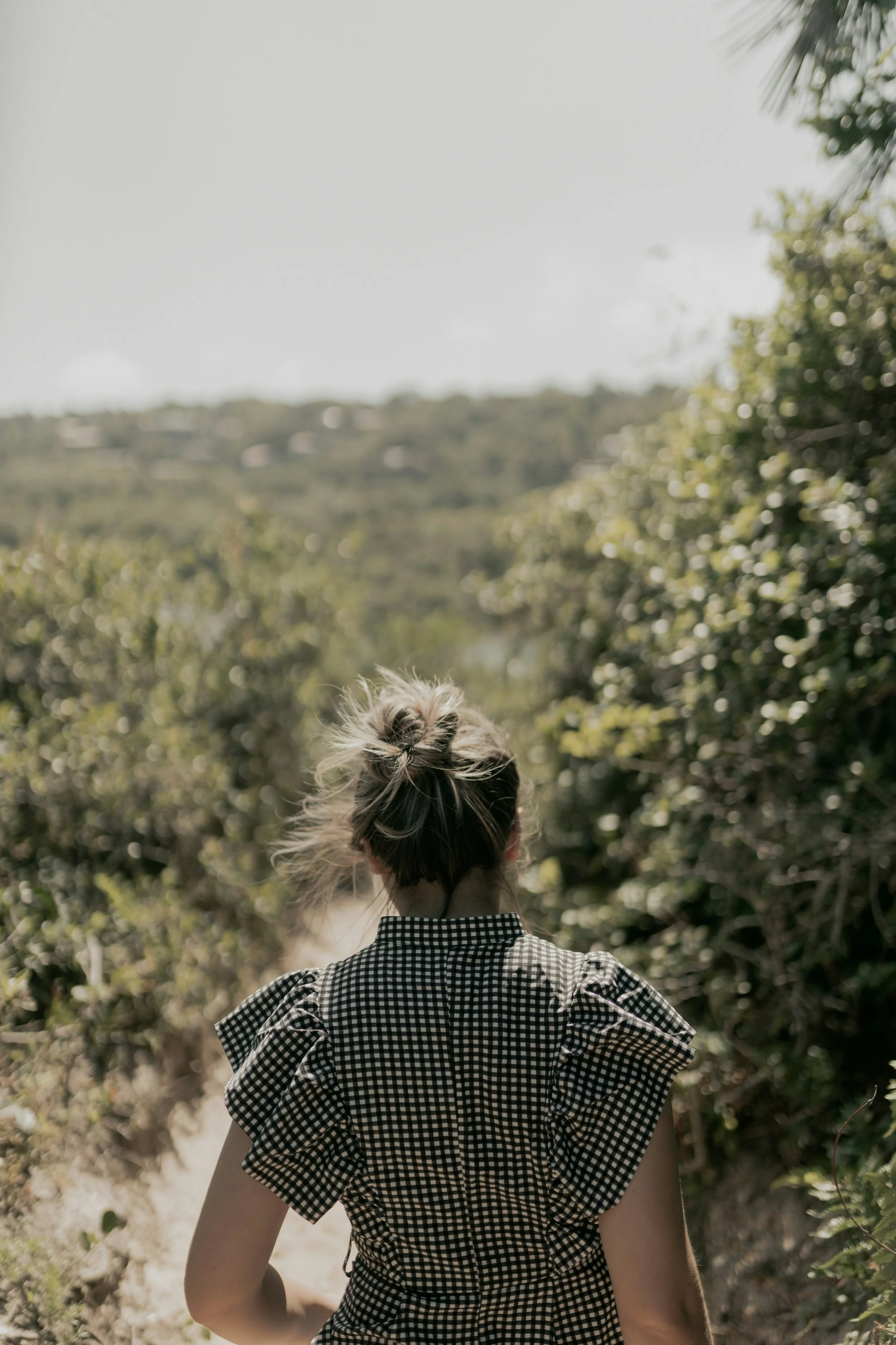 A person with blonde hair tied in a messy bun, wearing a black and white checkered dress, standing outdoors among greenery with trees and foliage, facing away from the camera.