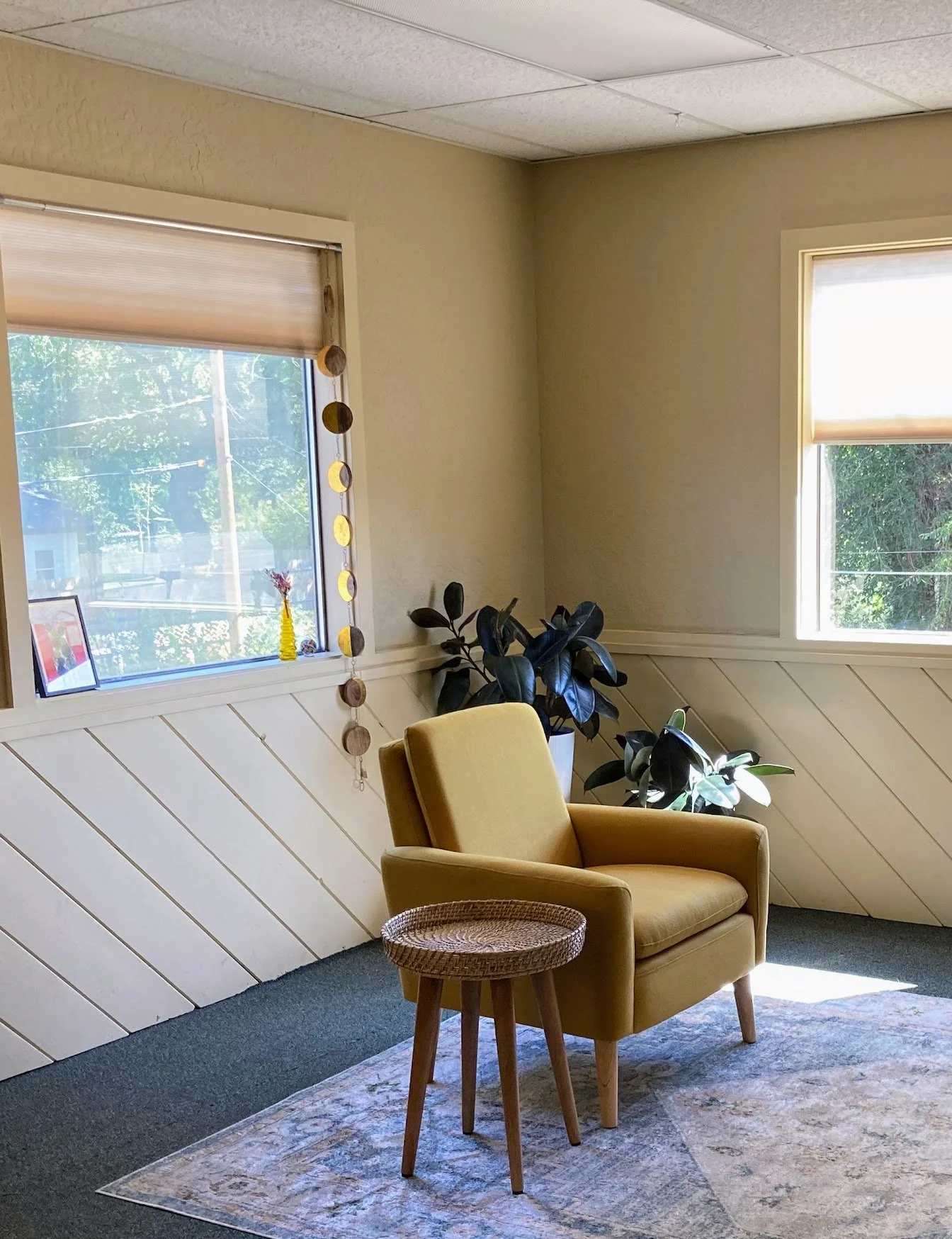 A cozy corner with a yellow armchair, a small woven side table, potted plants, and two windows with beige blinds allowing natural light.