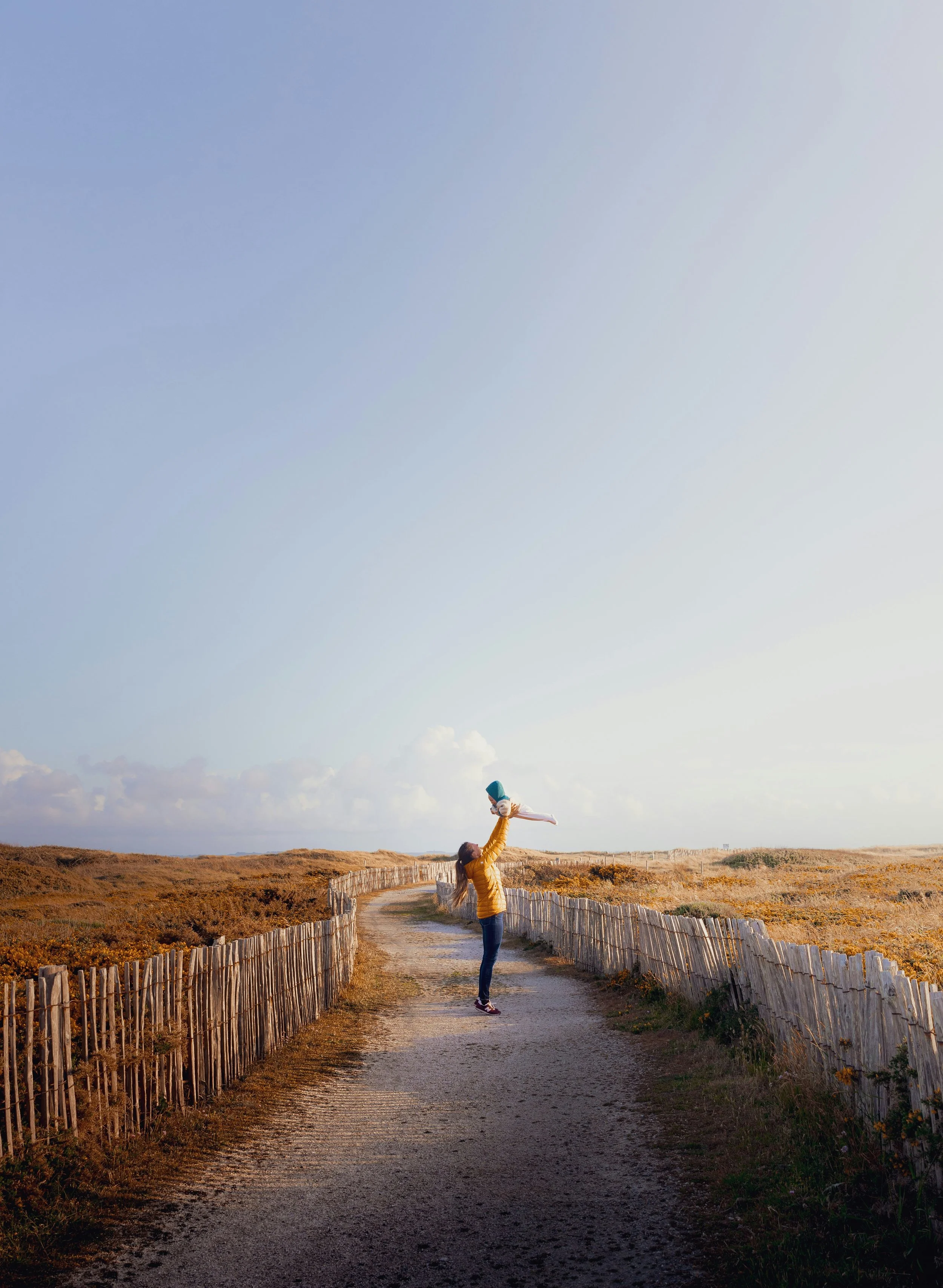 A woman lifting her child into the air on a dirt path in a rural landscape with fences and open fields during sunset.