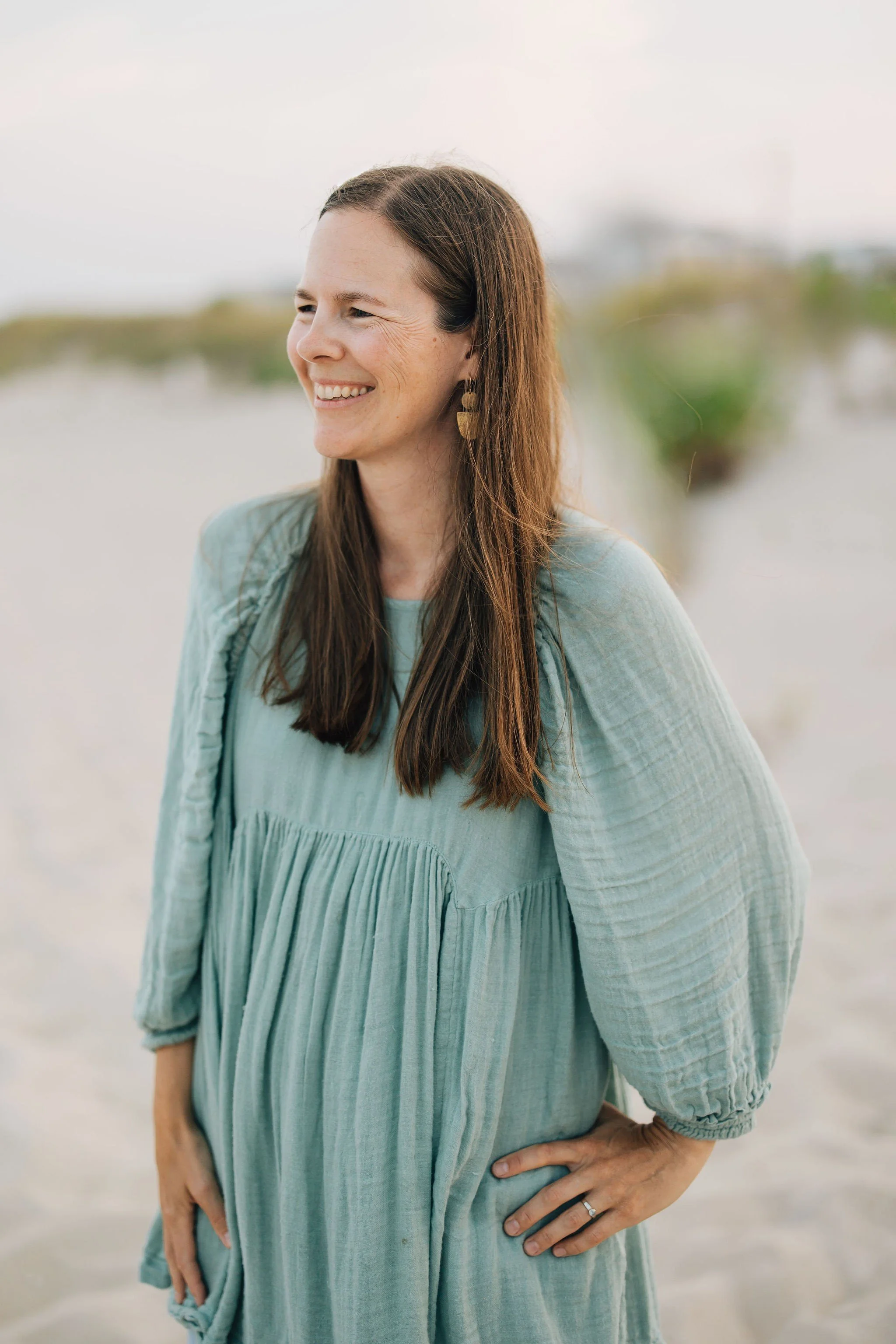 A woman with long brown hair, smiling, standing outdoors on a beach, wearing a loose, light blue dress with long sleeves and a ring on her left hand.