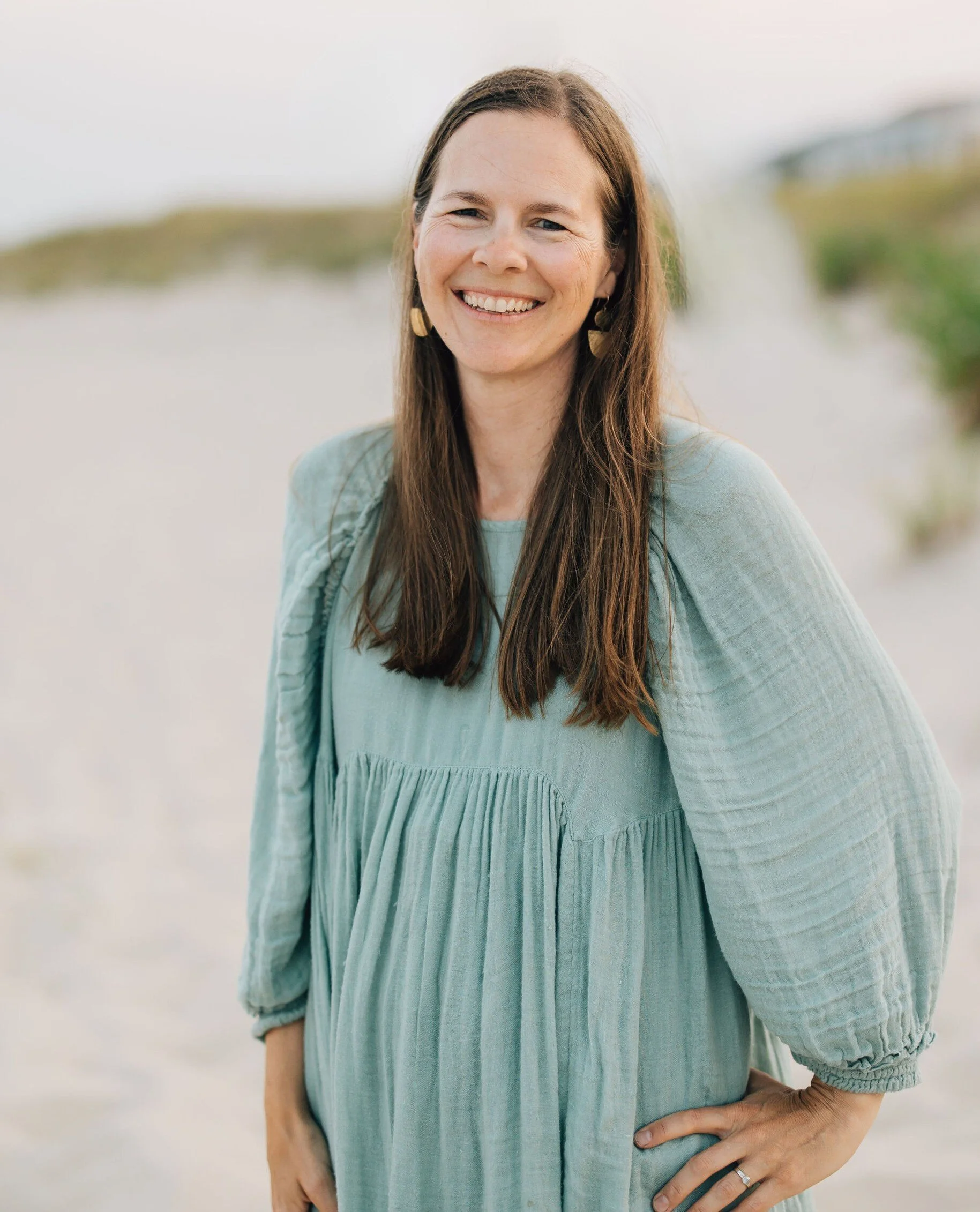 A woman with long brown hair, smiling, wearing a light blue dress and earrings, standing outdoors on a sandy area with green foliage in the background.