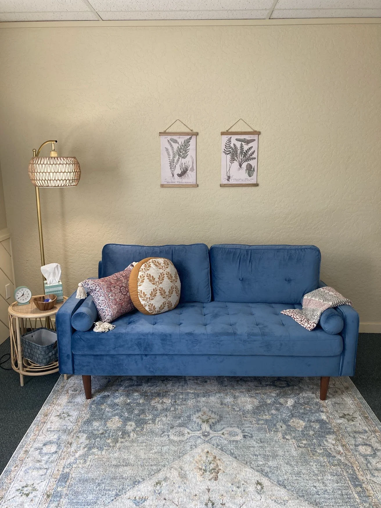 A blue velvet sofa with decorative pillows, a side table with tissues, a lamp, and wall art of fern illustrations.