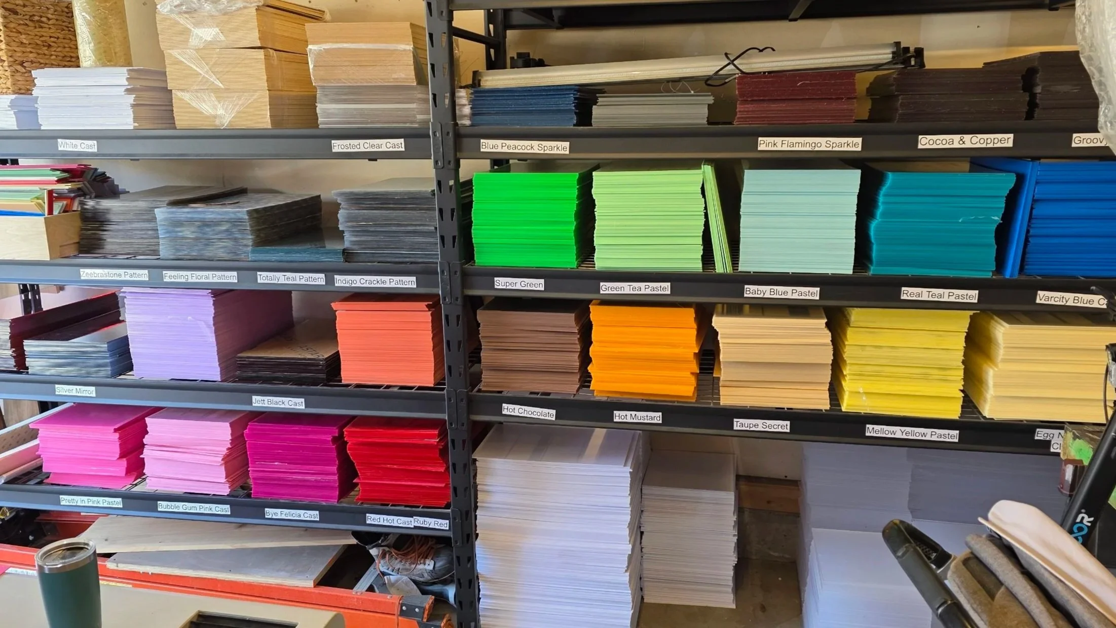 Shelves stocked with colorful paper sheets labeled with various patterns and colors, such as 'Frosted Clear Cast,' 'Blue Peacock Sparkle,' and 'Pink Flamingo Sparkle.'