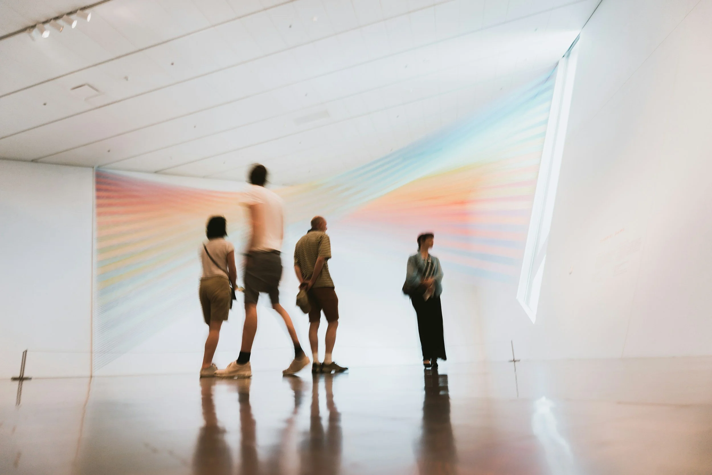 Four people standing in an art gallery, looking at a large, colorful, abstract artwork on the wall, with a skylight window illuminating the space.