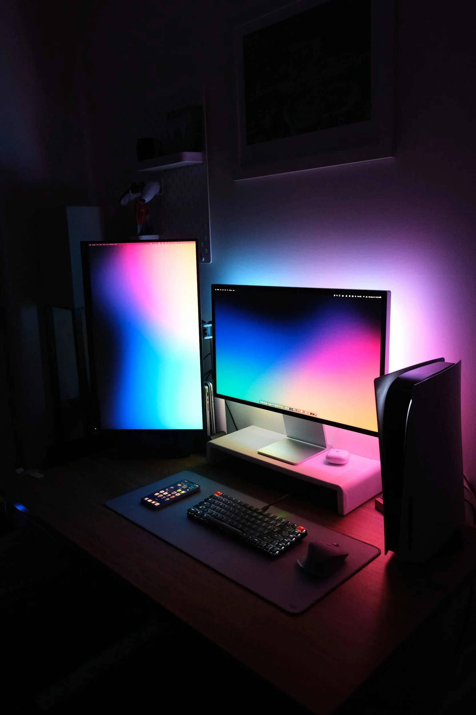 A computer desk setup with dual monitors displaying colorful gradient backgrounds, a smartphone, a mechanical keyboard with red accents, a mouse, and a pair of speakers, illuminated by colorful RGB lighting.