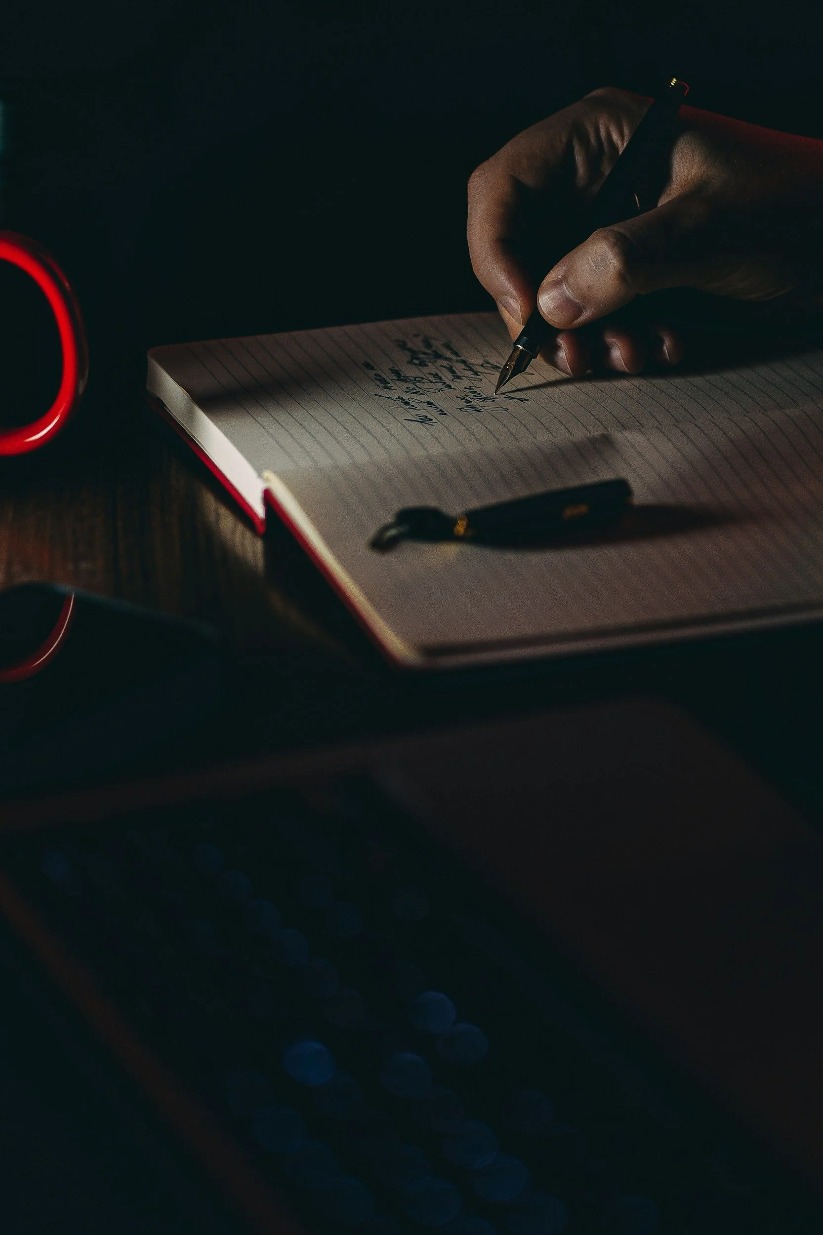 A person writing in a notebook with a black pen, sitting at a desk with a red mug and a black pen.