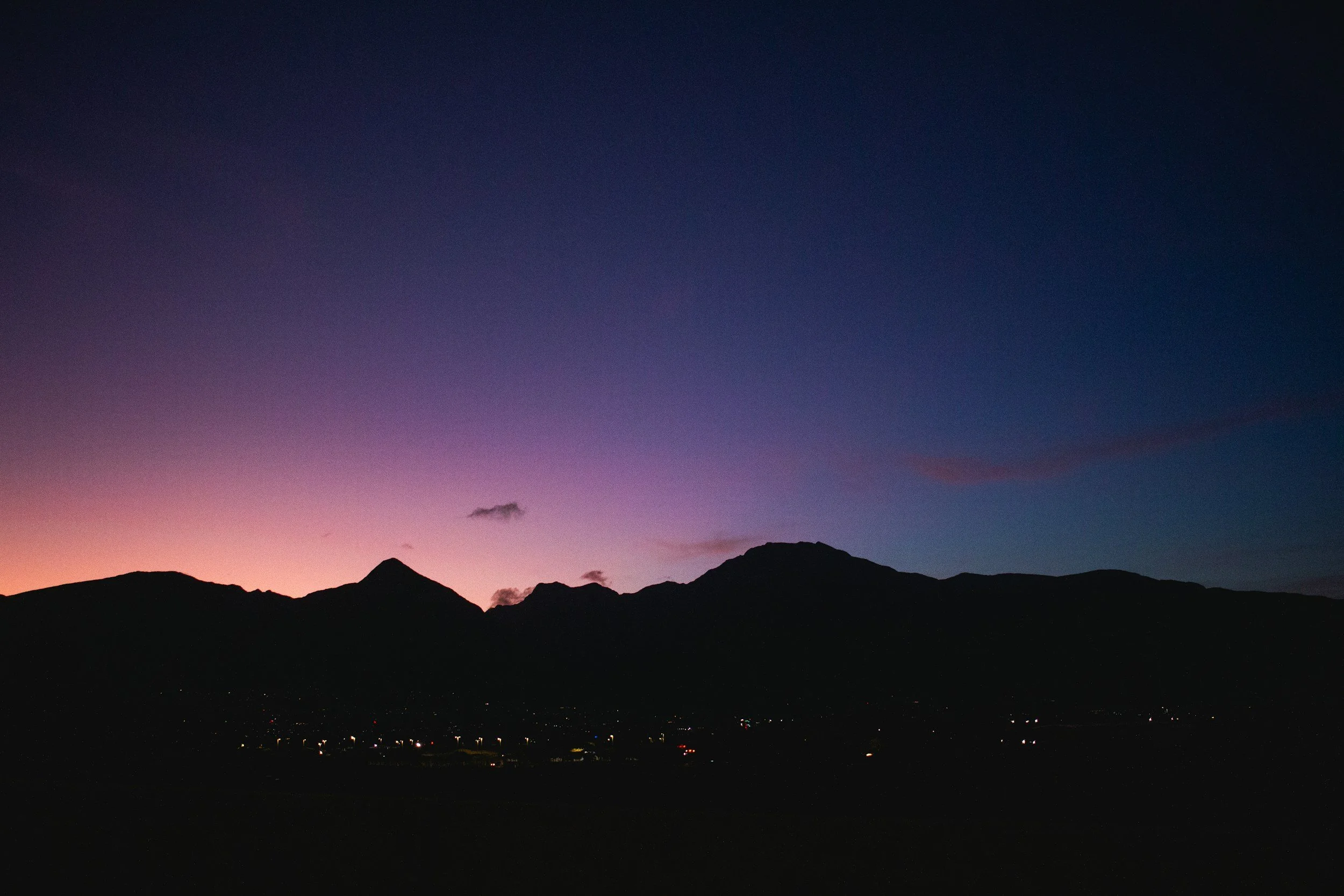 Silhouette of mountains during twilight with a colorful purple and pink sky and small lights at the base of the mountains.