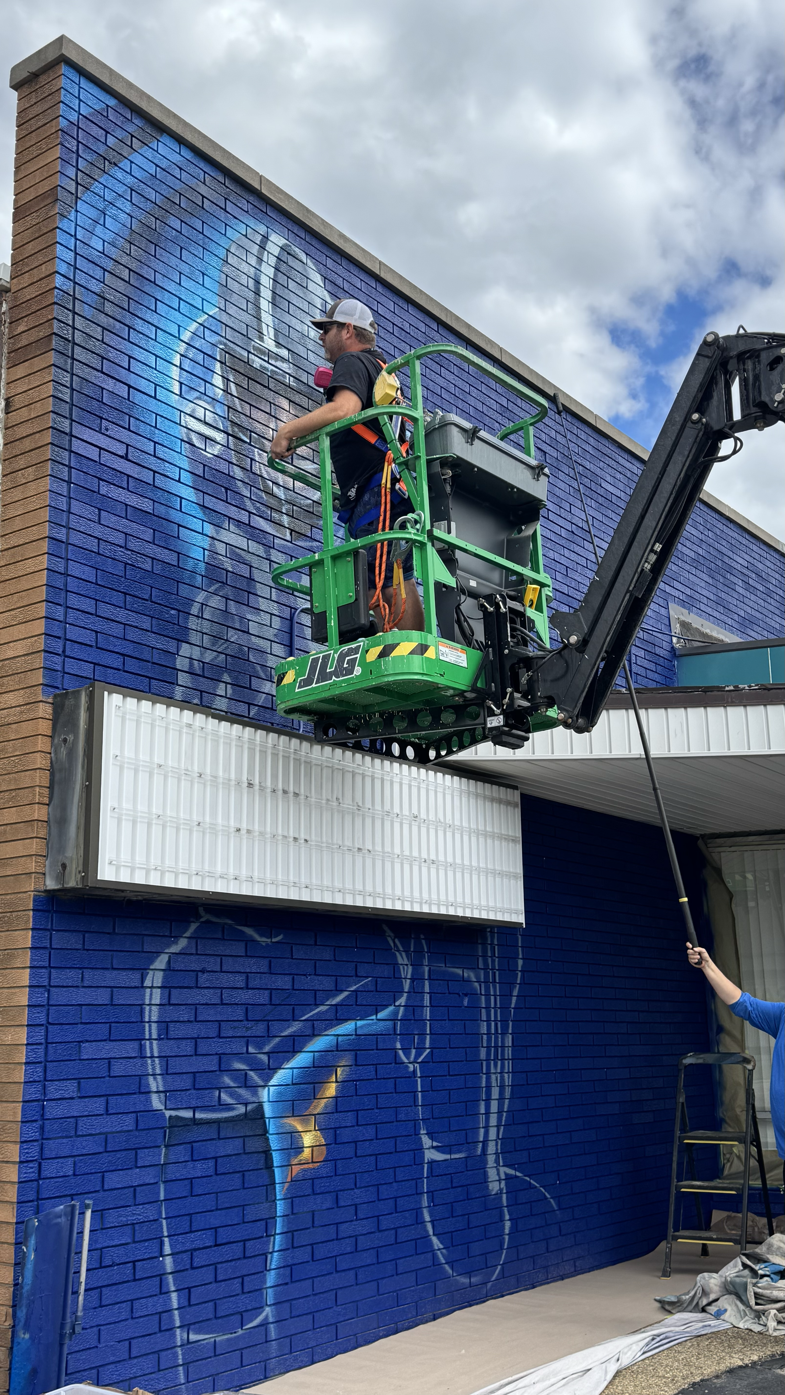 A man on a green lift painting a mural of a person's face on a blue brick wall outside a building. Another person is holding a long paint roller near the bottom right of the image.