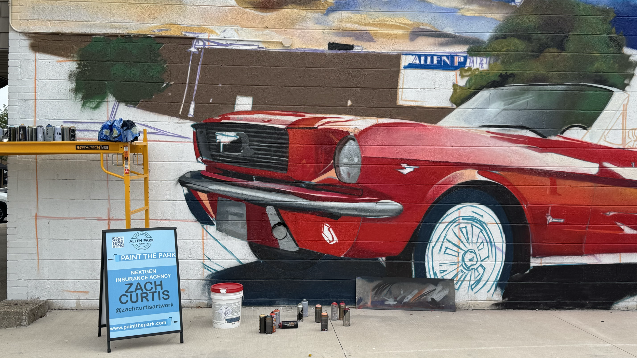 A mural of a red vintage Ford Mustang car being painted on a white brick wall. Paint cans and supplies are placed at the foot of the wall, and an A-frame signboard displays information about a community art event.