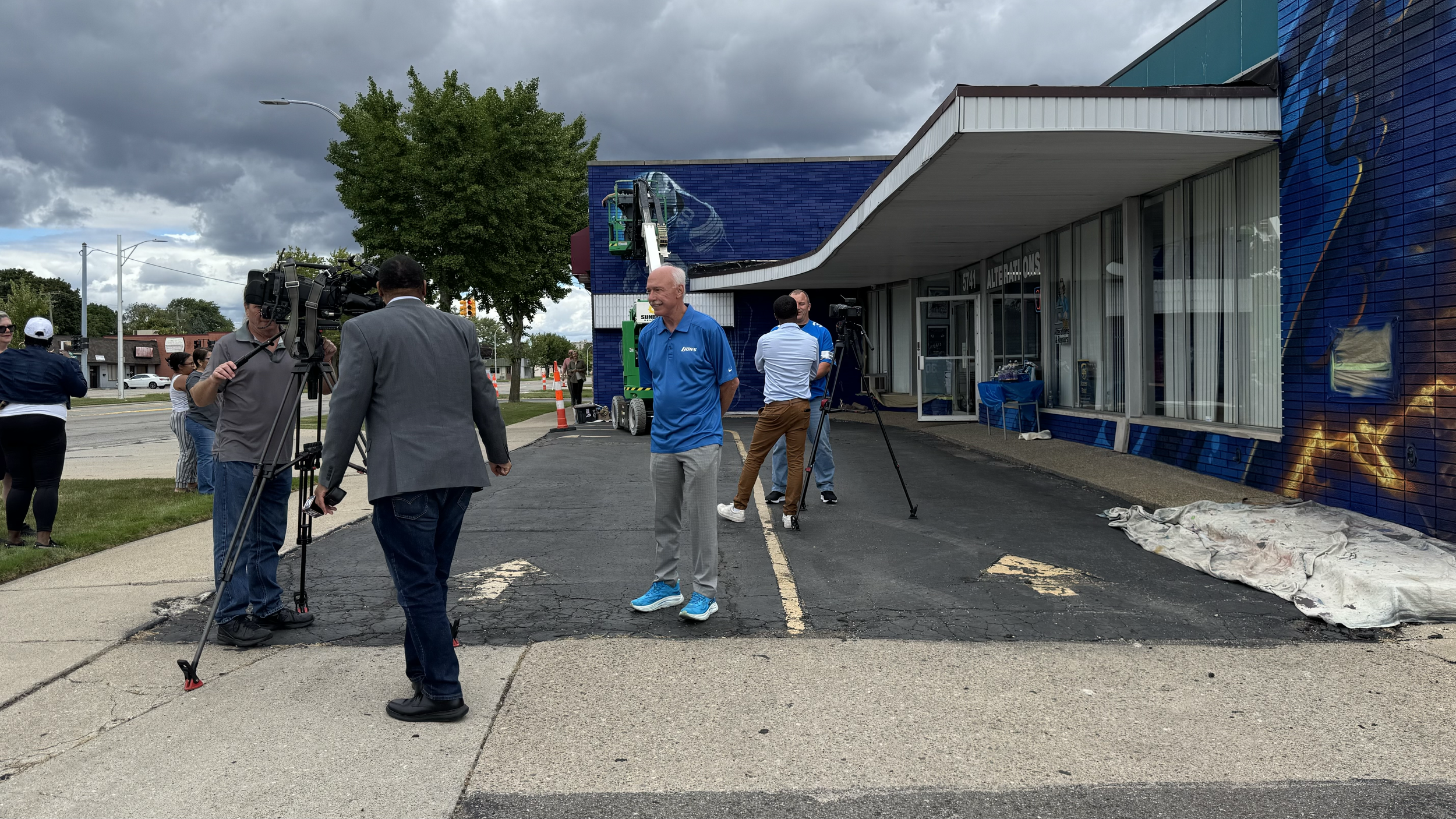 Group of people, including news crew with cameras, gathered outside a building with a mural on the wall and an overcast sky overhead.