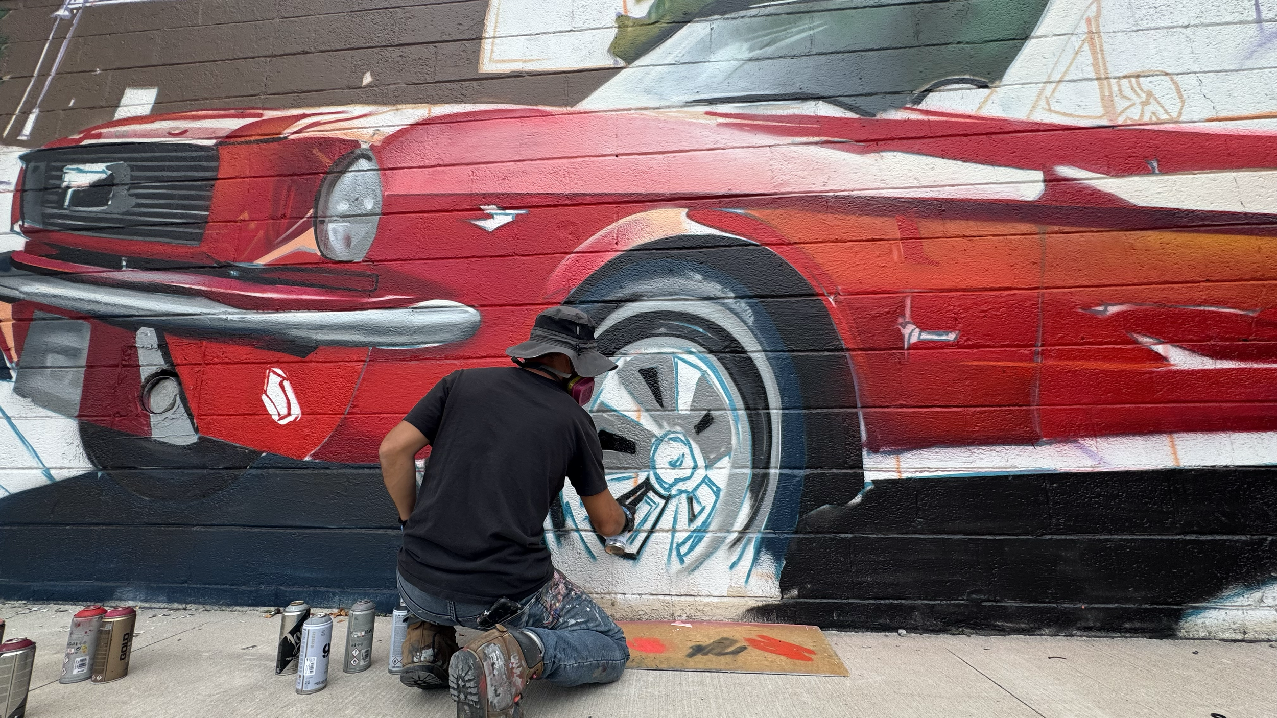 An artist painting a mural of a red vintage car on a brick wall. The artist is kneeling, wearing a black shirt, a bucket hat, and a mask, surrounded by spray paint cans.