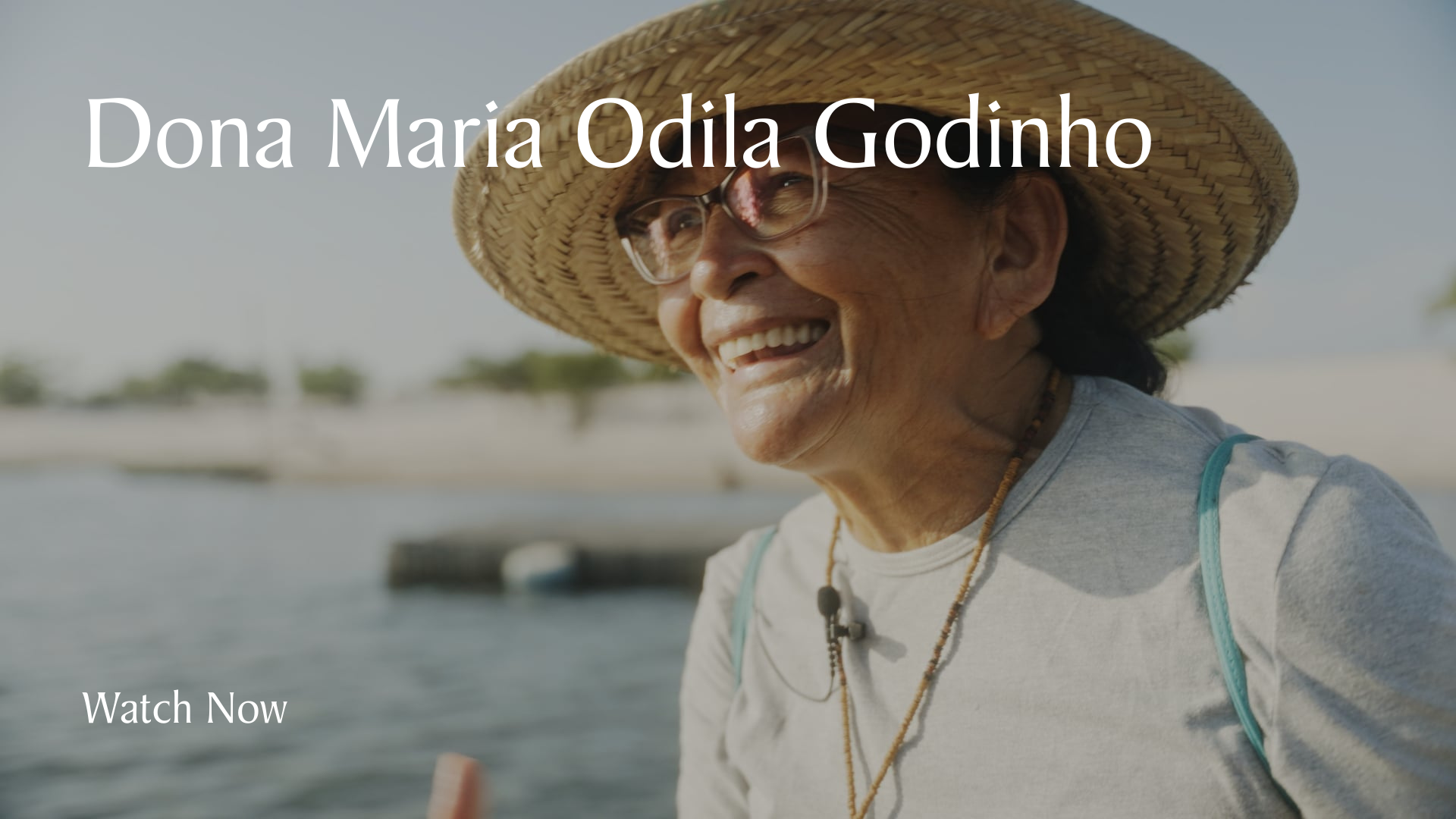 Dona Maria Odila Godinho. Smiling elderly woman wearing a wide-brimmed straw hat and glasses, outdoors near water with a blurred background of trees and sky.