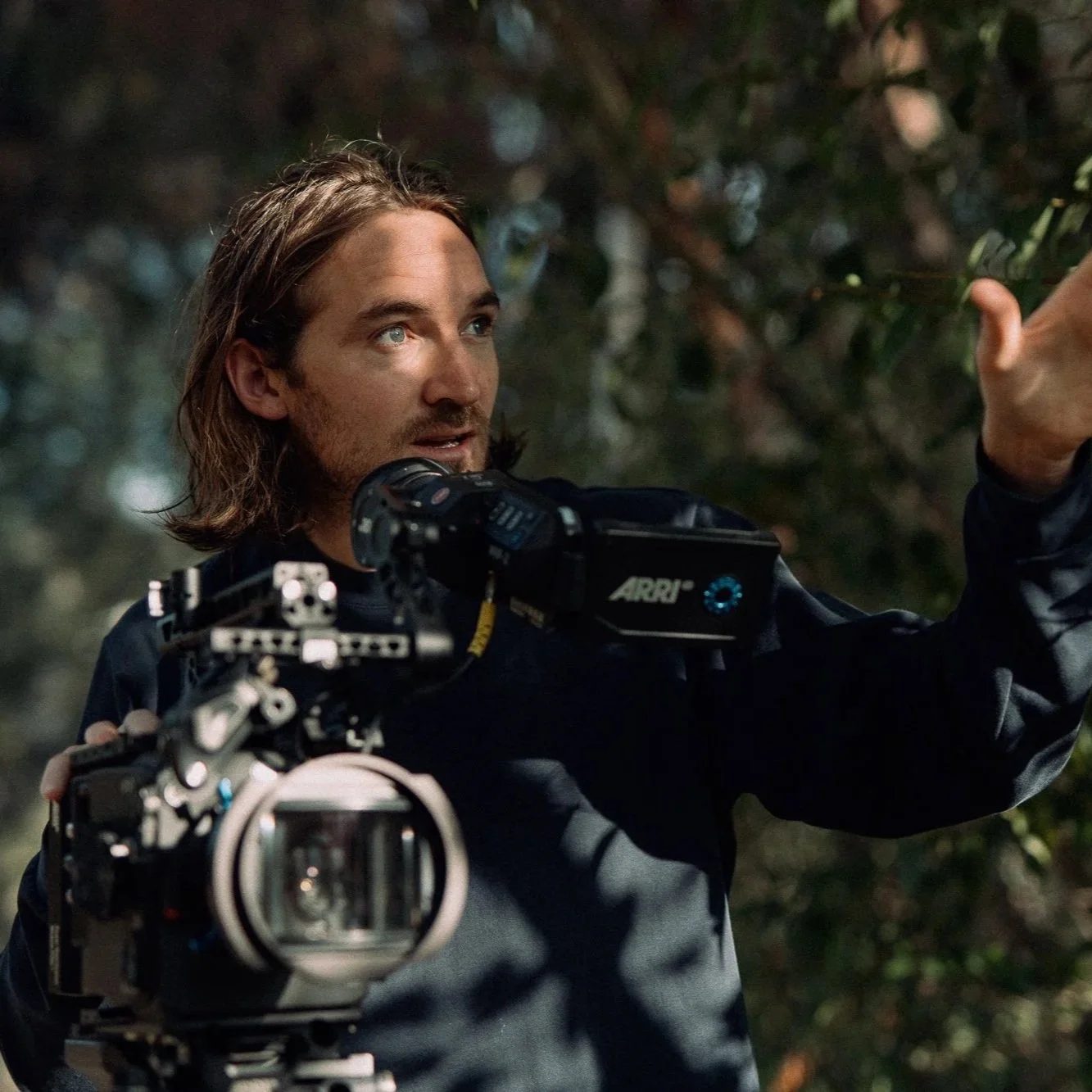A man with long hair and a beard, wearing a dark jacket, is holding a professional camera with a large lens and is reaching towards a tree branch in an outdoor setting at night.
