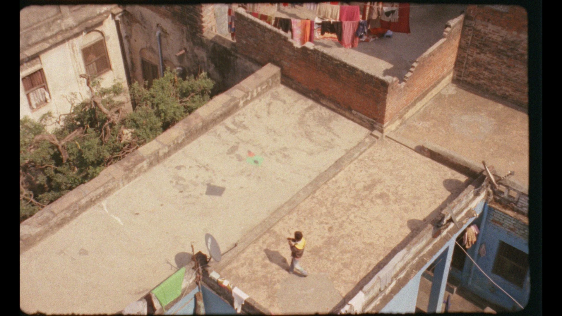 An aerial view of a rooftop in India with a person standing, laundry hanging on a clothesline, and clothes scattered on the rooftop surface, with neighboring buildings and trees visible.