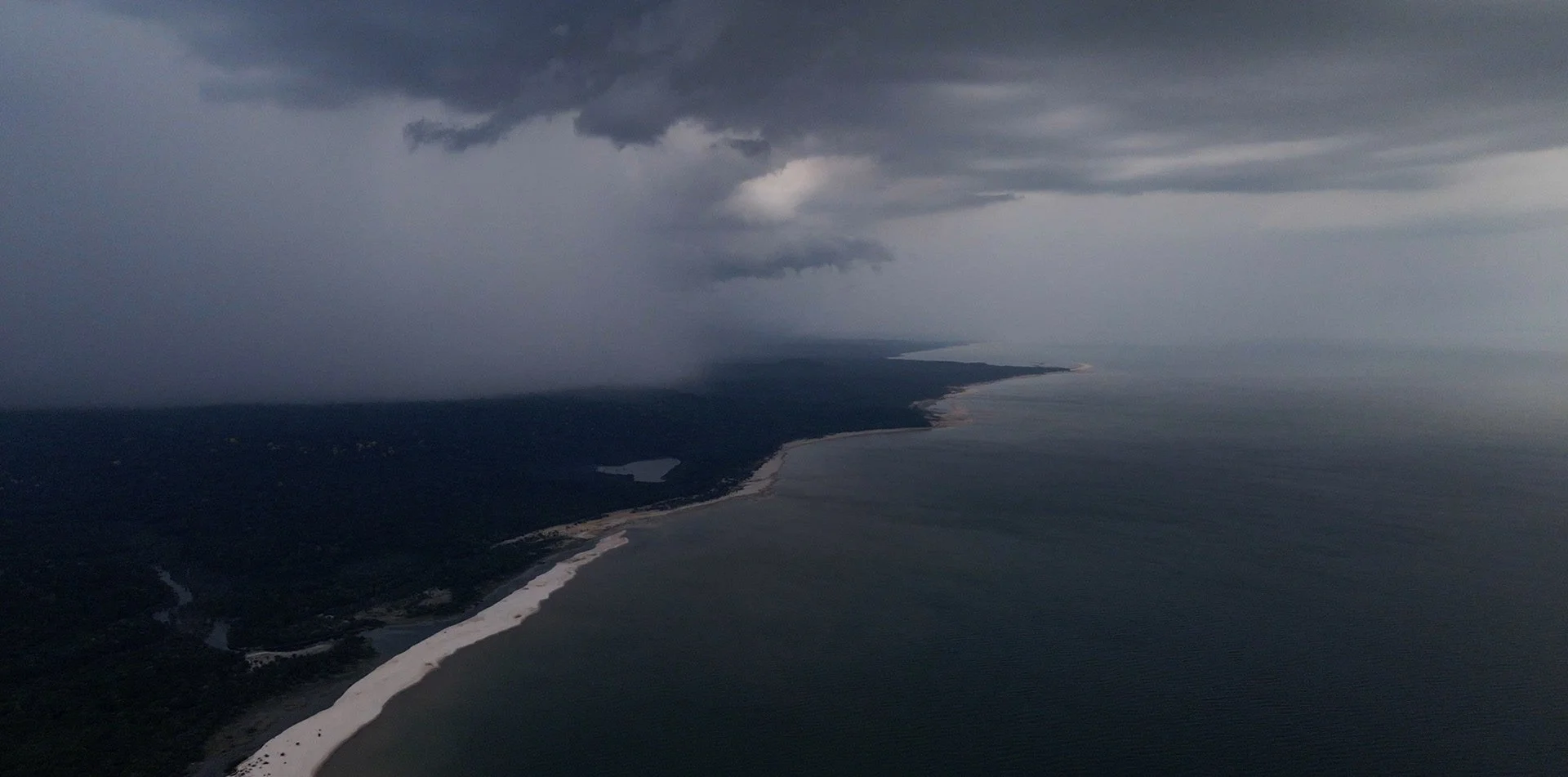 Aerial view of a coastline with dark storm clouds overhead, showing a sandy beach, lush green forest, and a dark ocean under a cloudy sky.