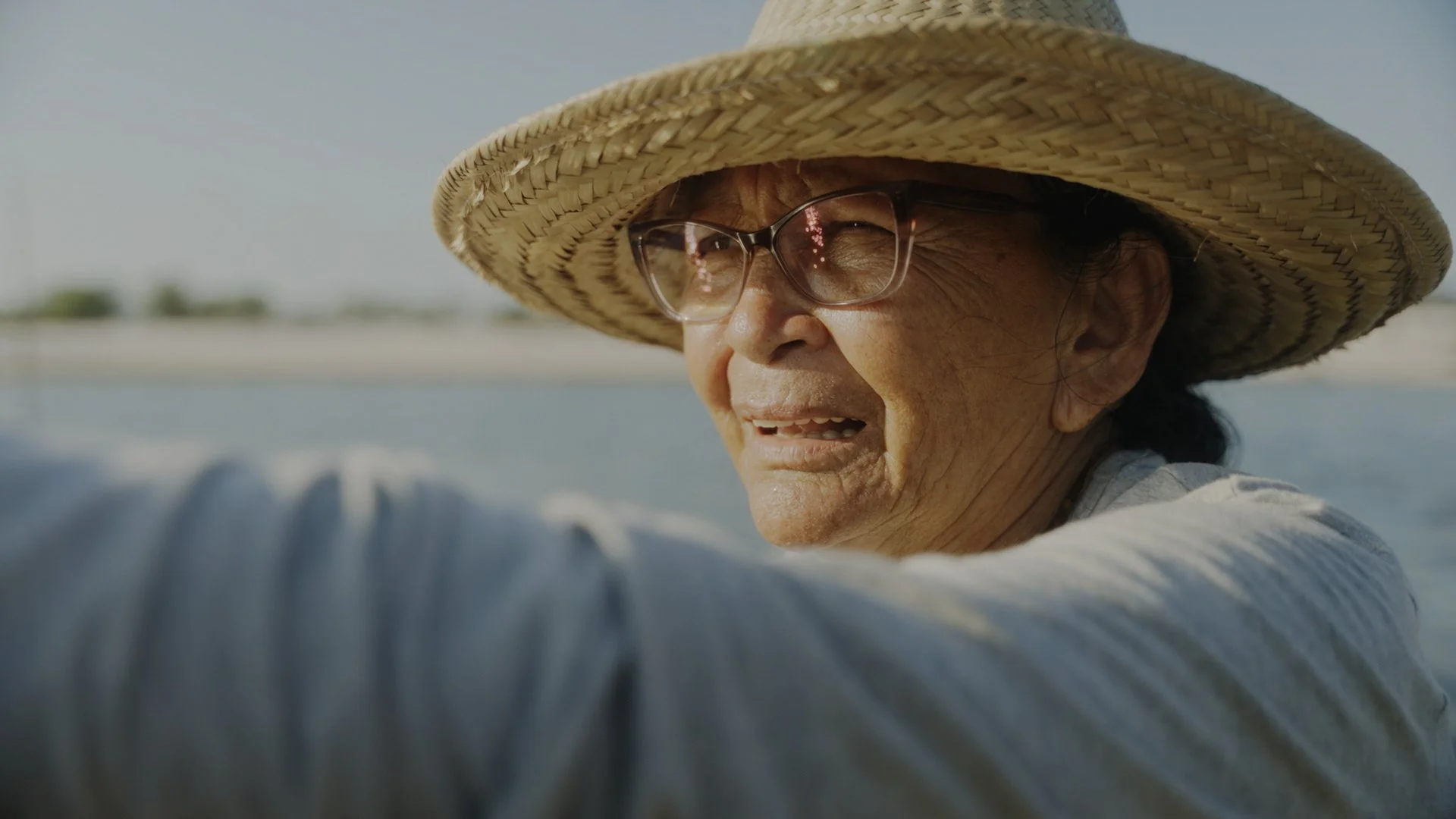 Close-up of an elderly woman in a large straw hat wearing glasses, making a playful face near a body of water with a distant shoreline.