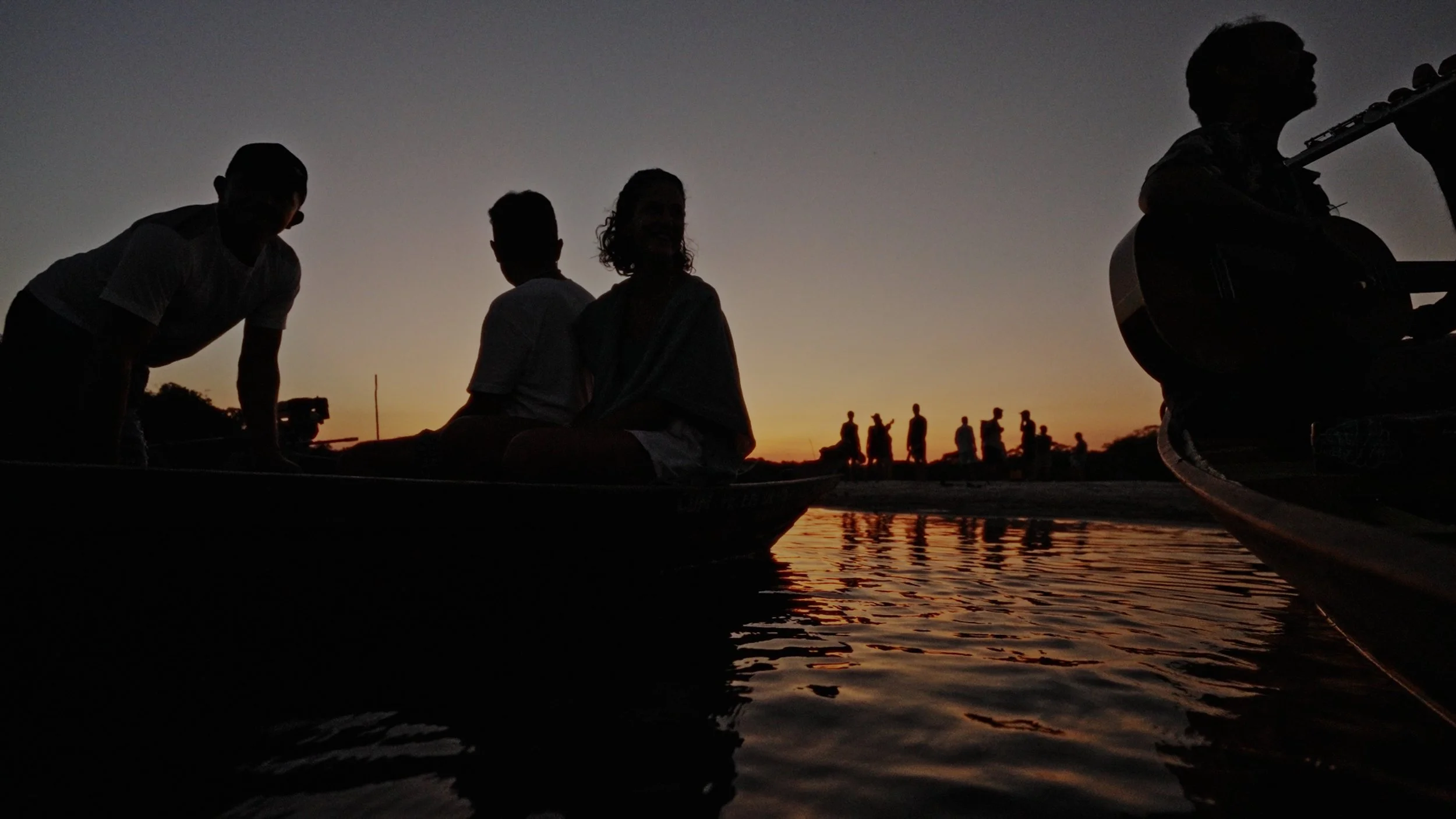 Silhouetted people near the water at sunset, with some sitting in boats and others walking along the shore.