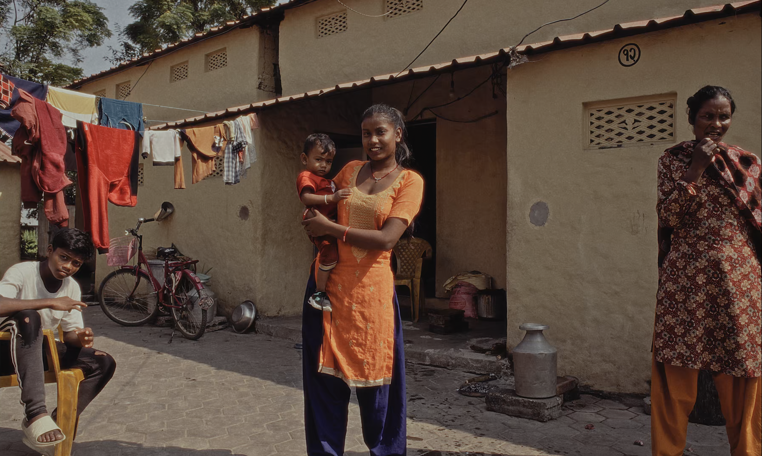 A woman in orange traditional attire holding a young child in red clothing stands outside a house with a group of people, including a boy sitting on a chair and another woman adjusting her scarf, with laundry hanging on a line and various household i