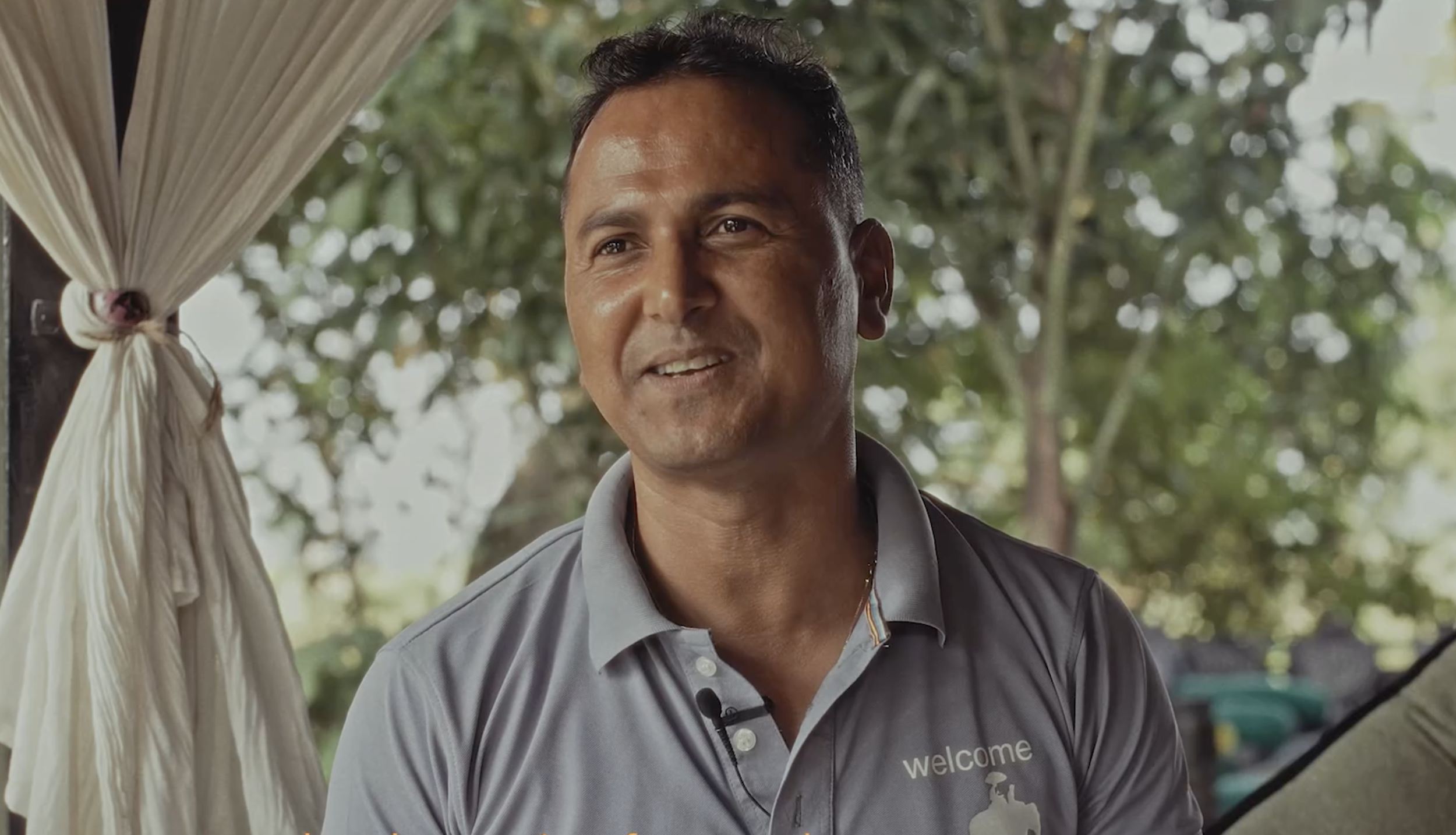 A man wearing a gray polo shirt with a palm tree logo and the word "welcome" embroidered on it, standing outdoors with trees in the background.