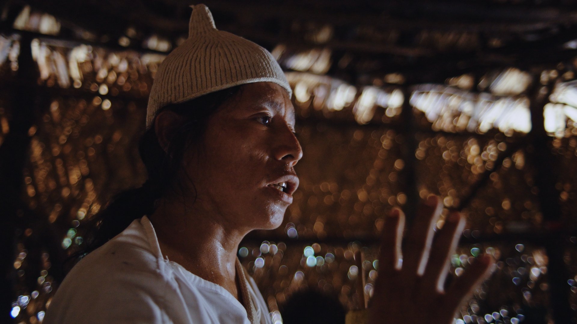 A tribe member wearing a beige knit cap, wearing a white shirt, speaking or explaining with their hand raised inside a structure with a thatched roof, with sunlight filtering through the thatch creating a bokeh effect in the background.