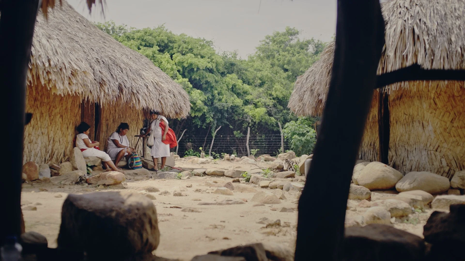 Four people in white attire sitting and standing outside between two traditional thatched-roof huts, with a rocky ground and green trees in the background.