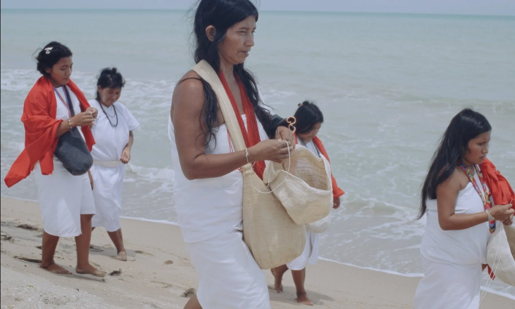 A group of women and children walking barefoot on a beach, wearing traditional white clothing with red and colorful accessories, carrying bags.