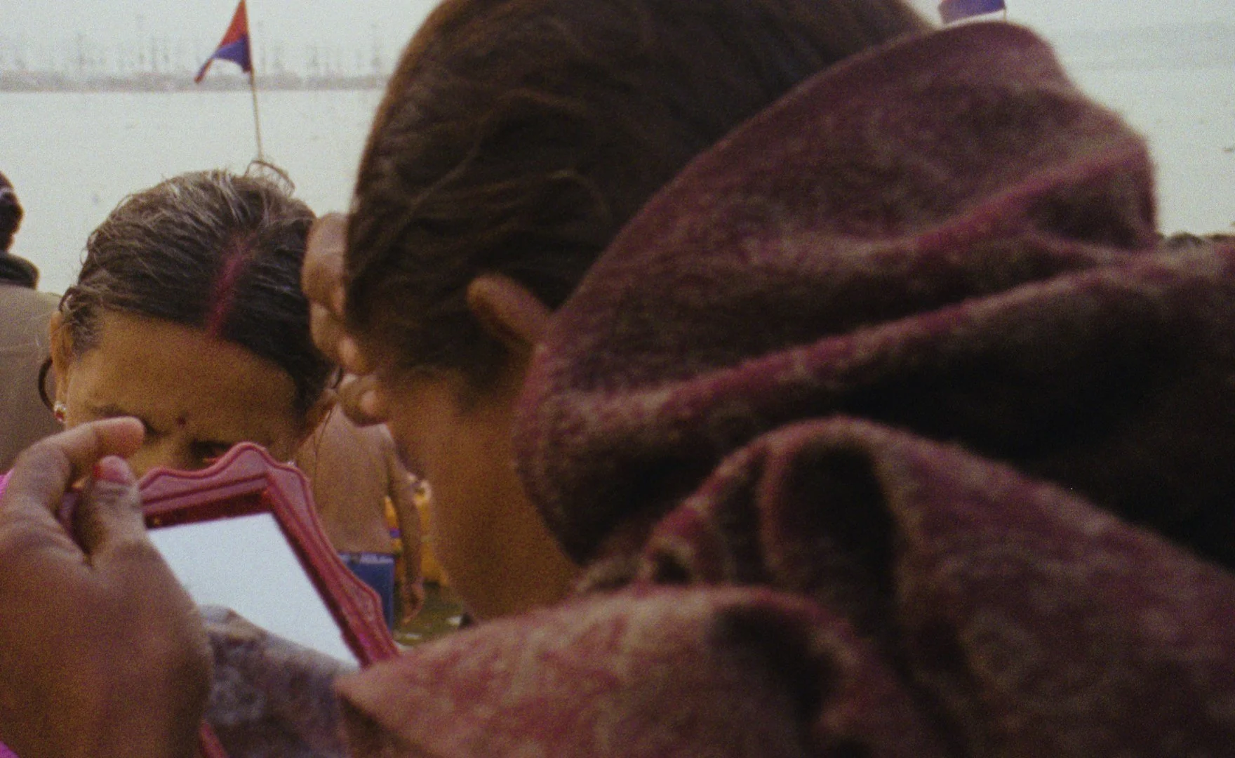 Close-up of two women with braided hair; one is looking at a phone; in the background, a flag and multiple people are visible.