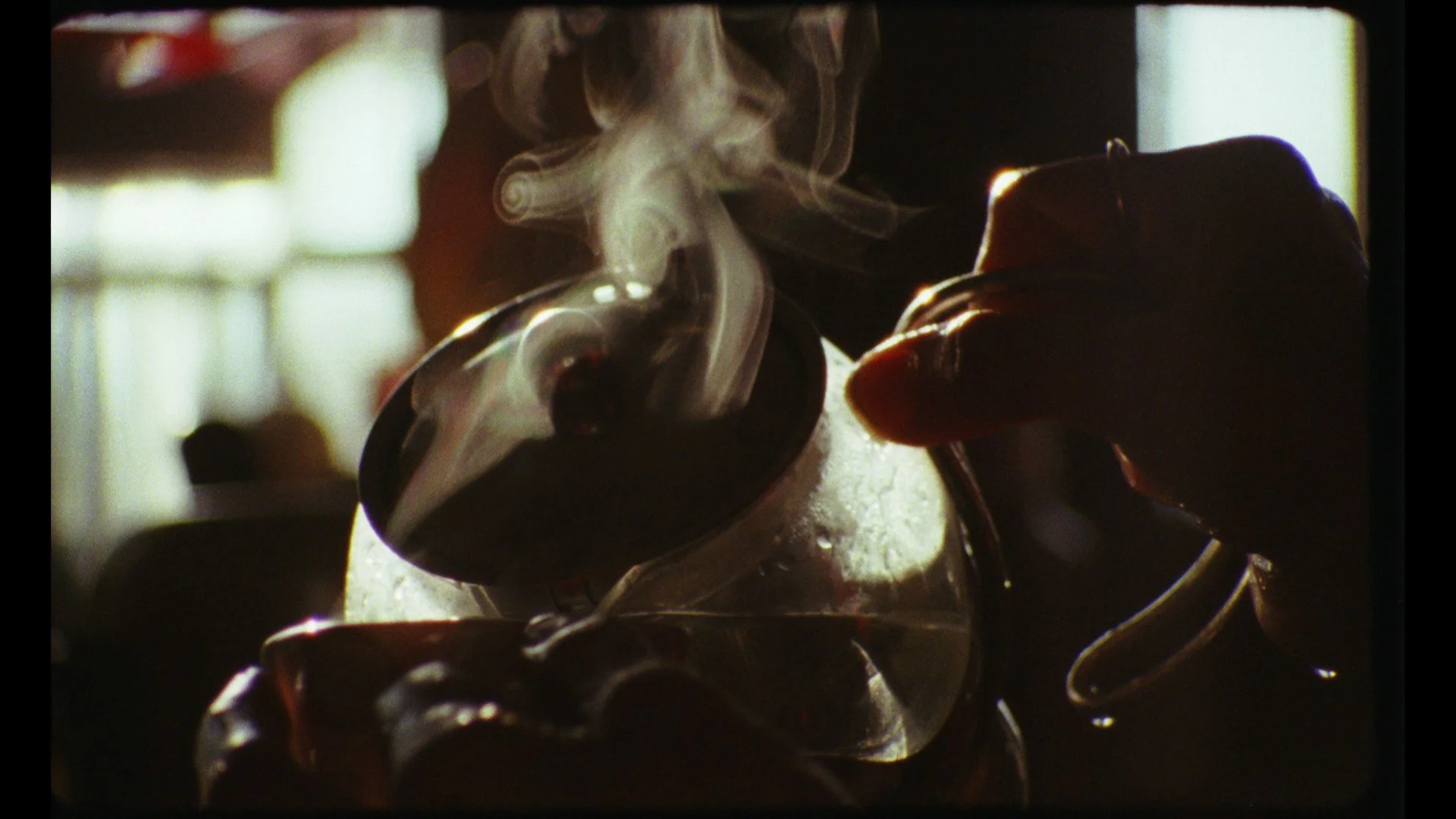 Close-up of a hand holding a glass cup with a hot beverage, with steam rising from it, against a blurred background.