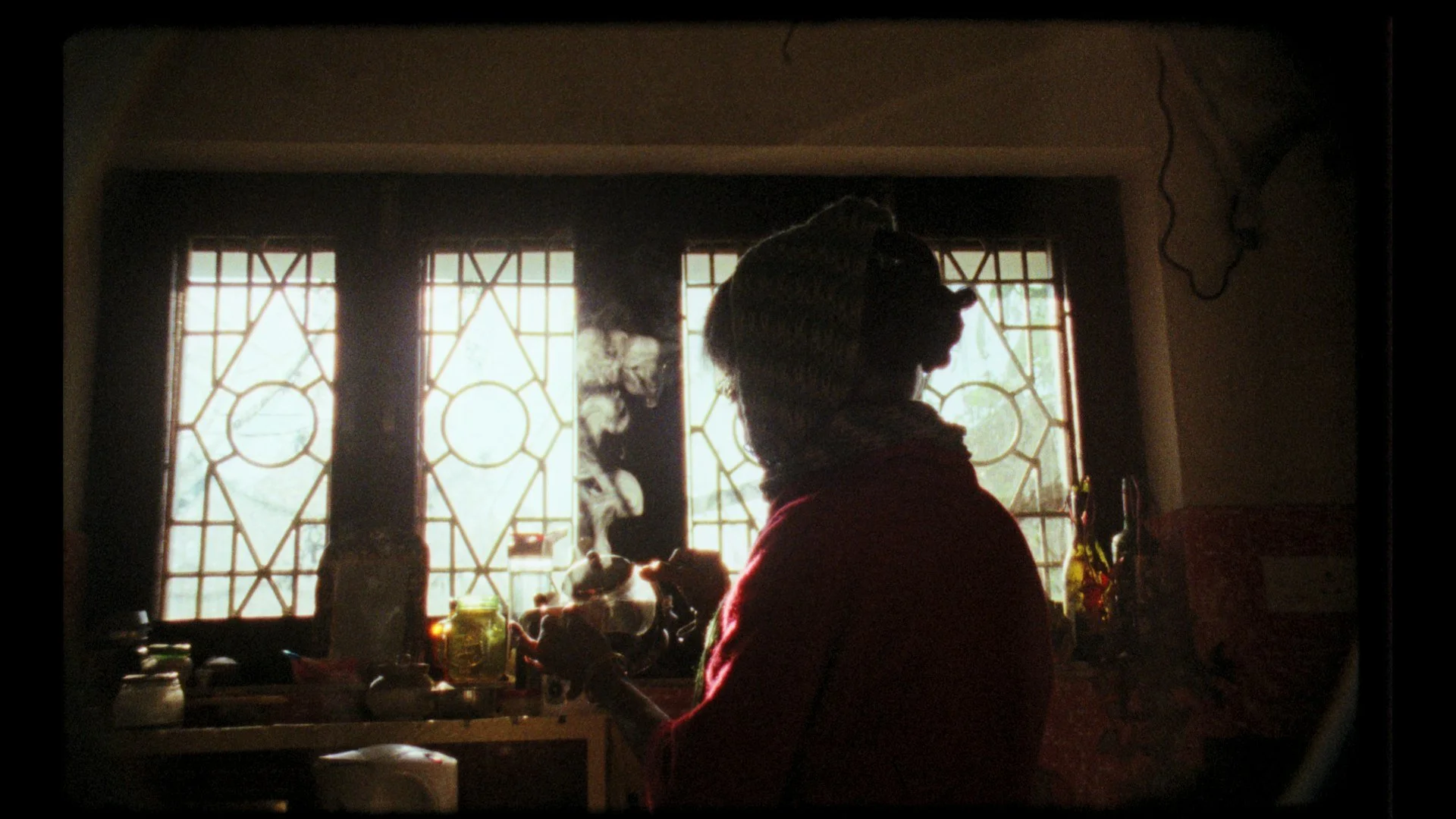 A silhouette of a woman with a bun hairstyle, in a kitchen, smoking while standing in front of a window with decorative bars, and various kitchen items on the counter.