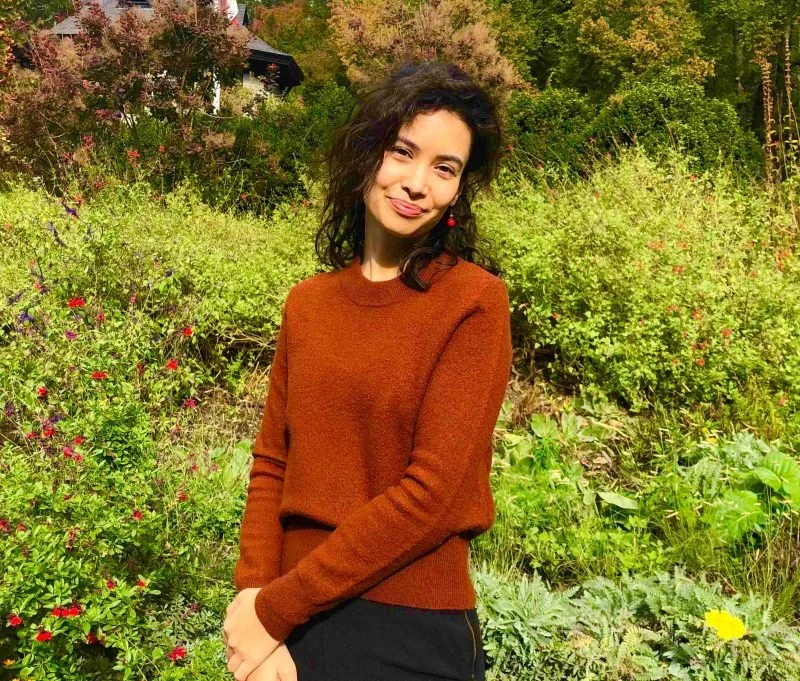 Young woman with curly dark hair wearing an orange sweater standing outdoors among green shrubs and flowering plants.