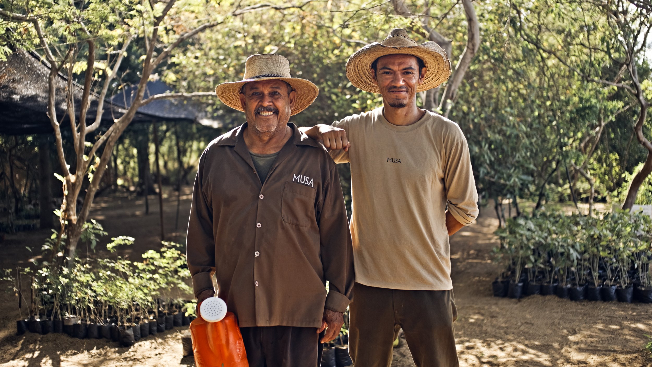 Two men wearing straw hats standing in a garden with young plants, one with his arm around the other's shoulder, both smiling.