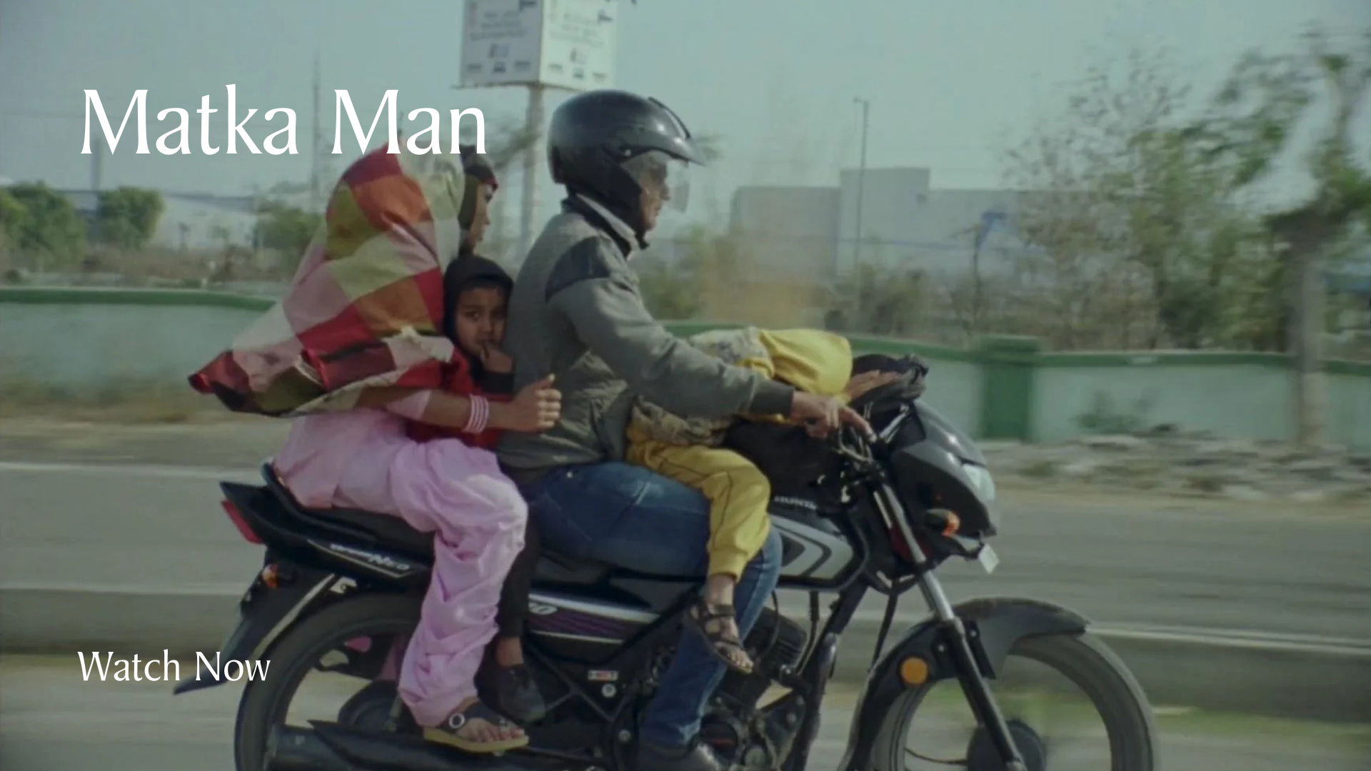 A man driving a motorcycle with his wife and two children, and they are traveling on a road with a cityscape in the background.