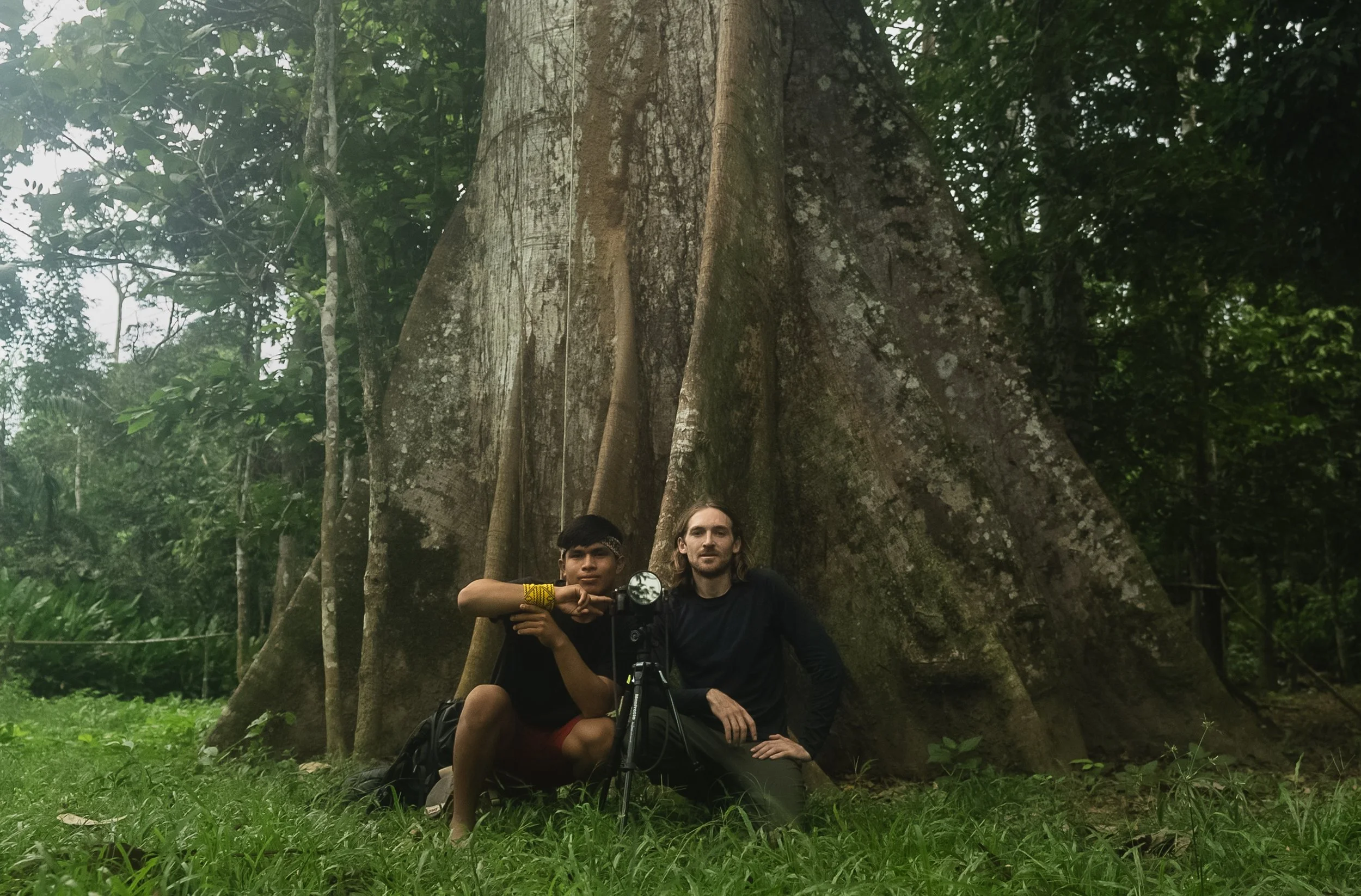 Two people sitting on grass in front of a large tree in a jungle, one with long hair and the other young with short dark hair, with camera equipment.