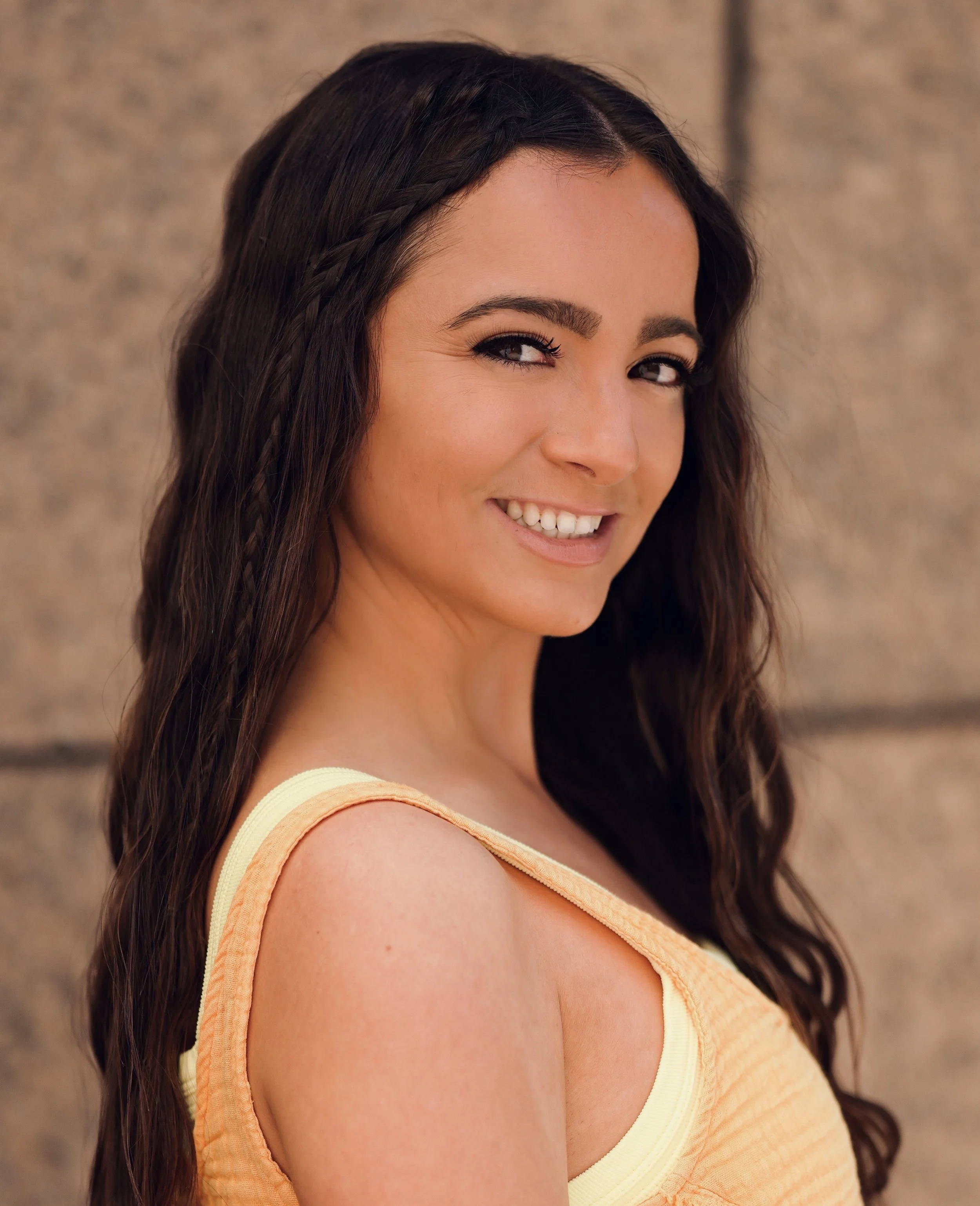 A woman with long wavy dark hair and a bright smile, wearing an orange sleeveless top, standing outdoors against a stone wall background.