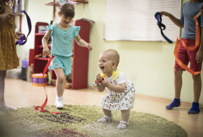 Children playing with ribbons on a carpet in a playroom, with a baby kneeling and smiling.