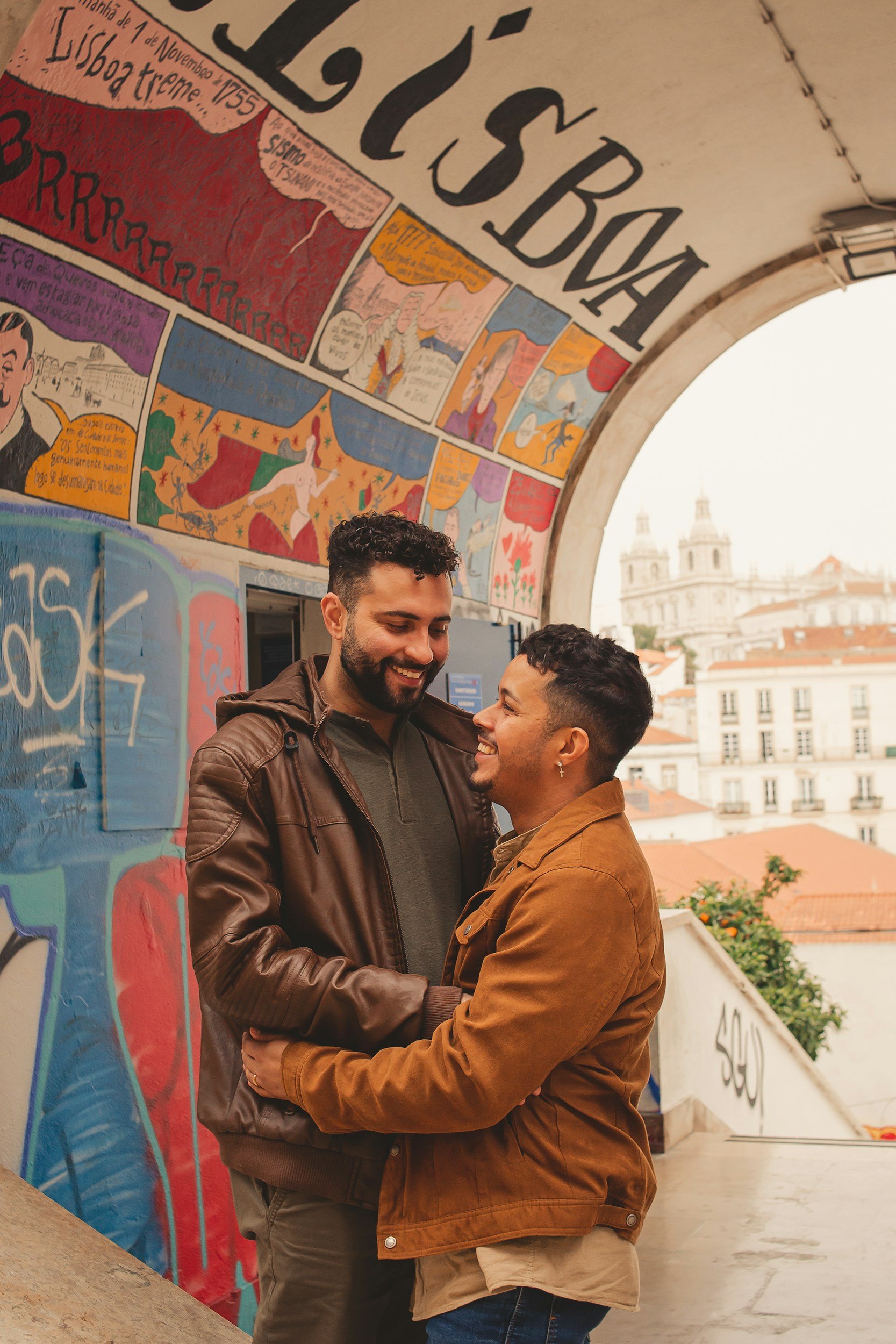 Two men smile and embrace under a colorful tiled mural on a city bridge, with historic buildings in the background.